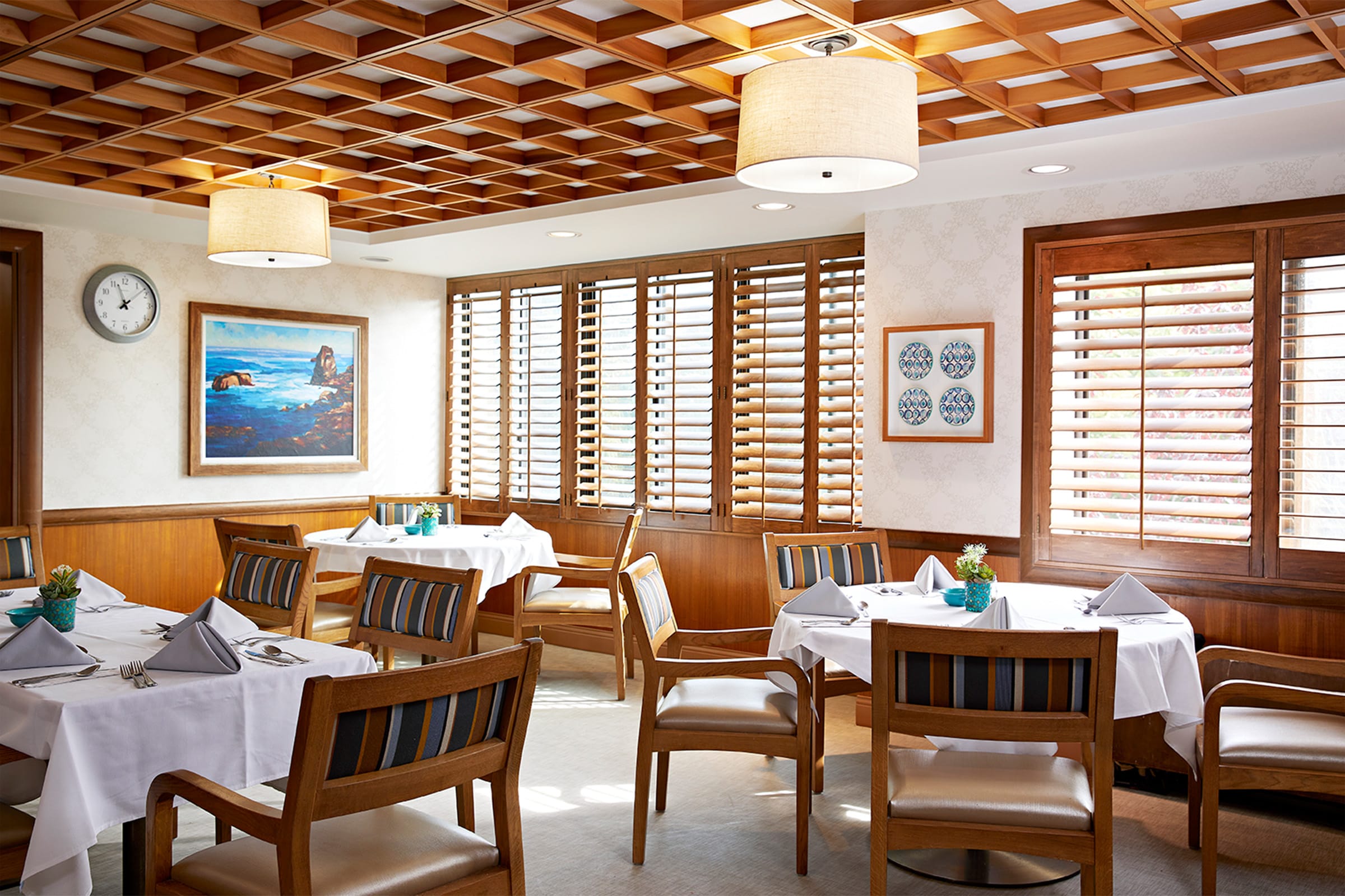 A bright, elegant restaurant dining area with wooden chairs and tables set with white cloths and folded napkins. Large windows with wooden shutters let in natural light, and artwork decorates the walls.