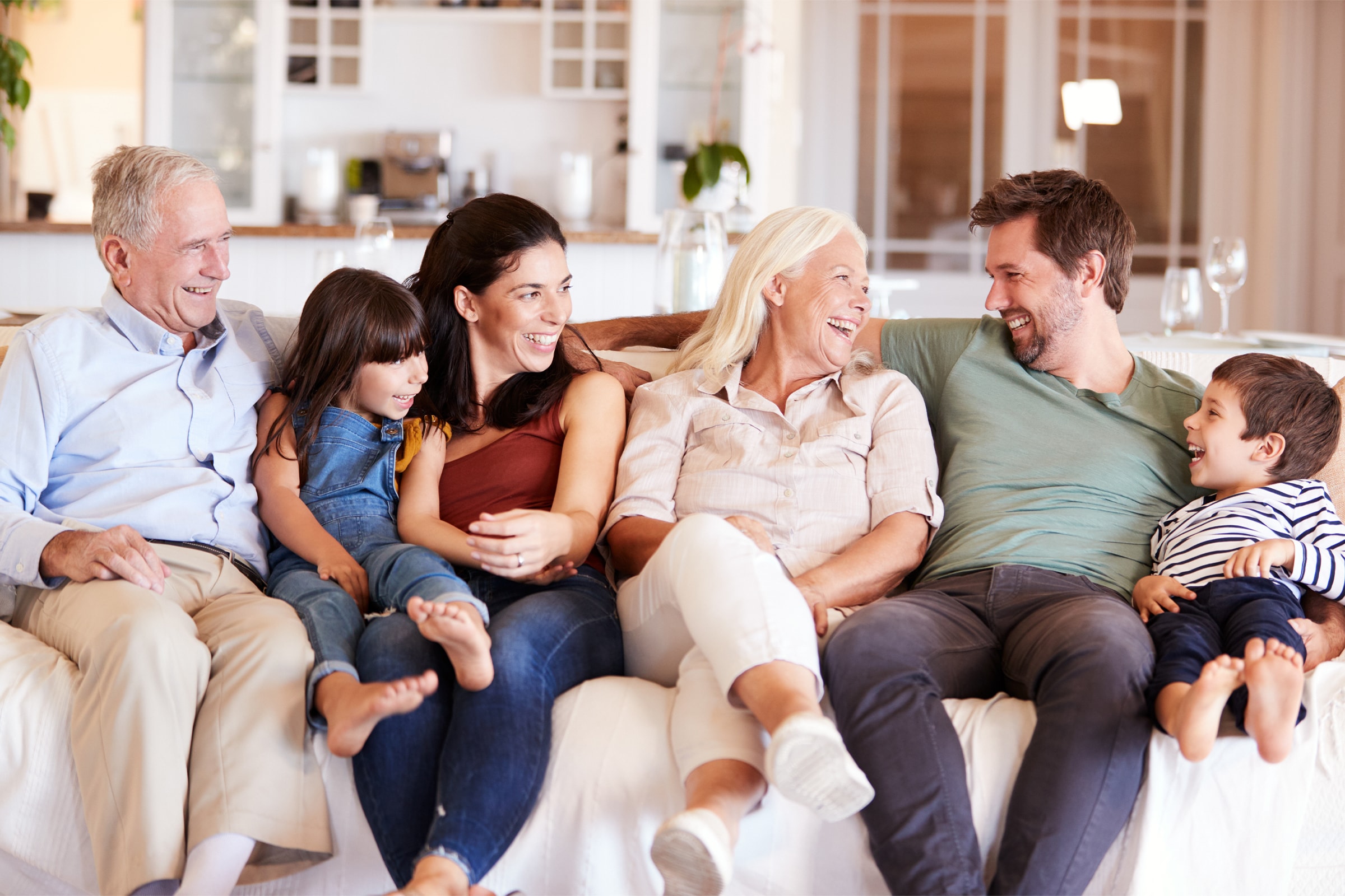 Three generations of a family sit together on a couch, smiling and laughing, with grandparents, parents, and two young children enjoying each others company in a cozy, bright living room.