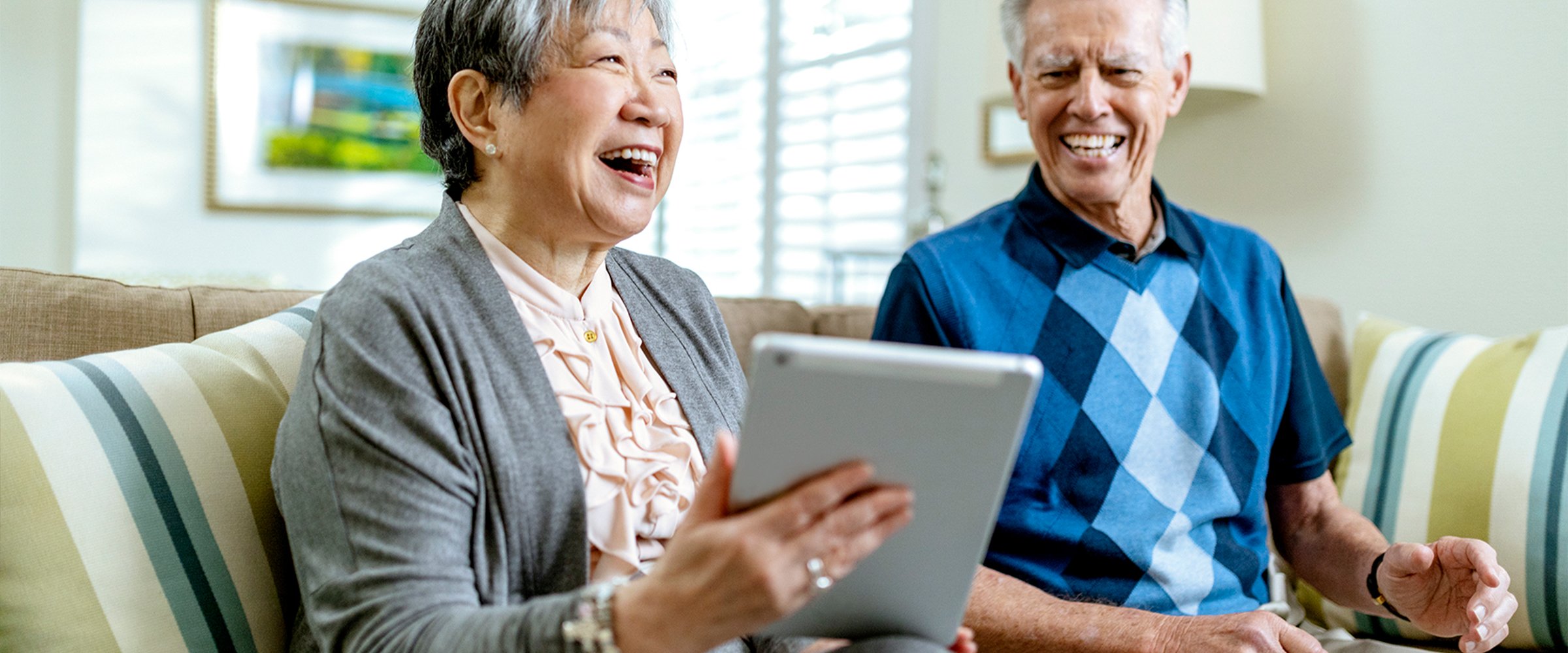 Two smiling older adults sit together on a sofa, holding a tablet and looking at the screen, appearing happy and engaged as they read a blog in a bright, cozy living room.