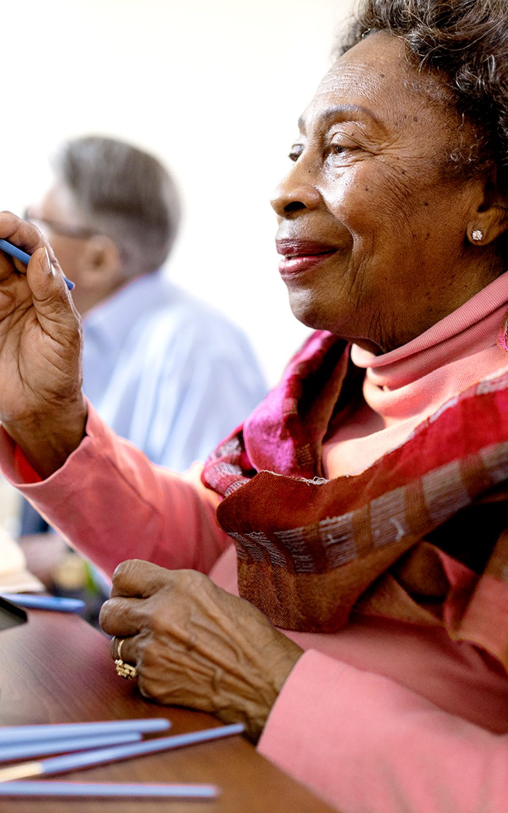 An elderly woman wearing a pink sweater and colorful scarf smiles while holding a blue pen, seated at a table. Another person is blurred in the background. The mood is warm and engaged.