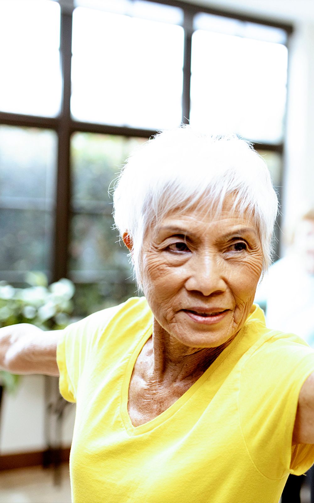 An older woman with short white hair wears a yellow shirt and stretches her arms out to the sides in a bright room, looking to her right and smiling slightly.