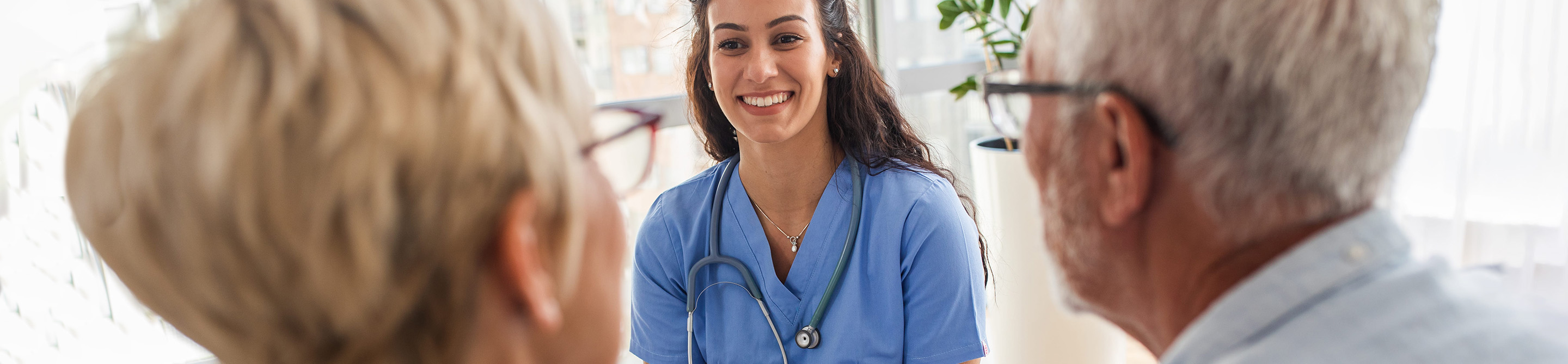 A smiling female healthcare professional in blue scrubs with a stethoscope speaks with two older adults in a bright, sunlit room.