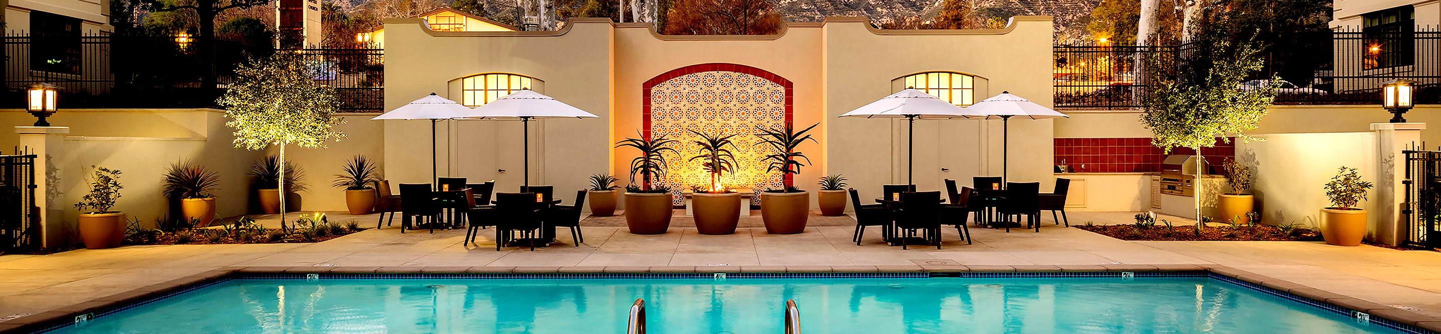 A rectangular outdoor pool with lounge chairs, tables, and white umbrellas surrounded by potted plants, warm lighting, and a decorative tiled wall in the background. Trees and mountains are visible behind a fence.