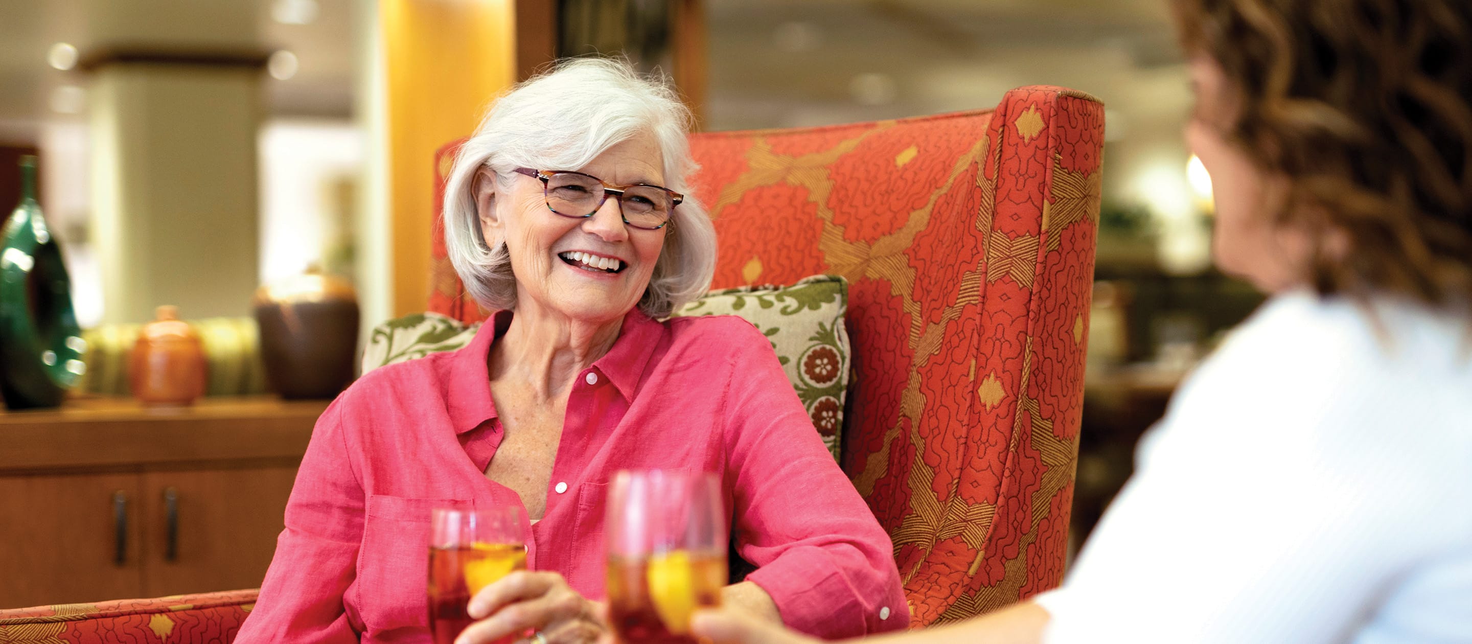 Hero An older woman with white hair and glasses, wearing a pink shirt, smiles while holding a drink and sitting in a patterned armchair. Another person holds a drink across from her in a cozy, warmly lit room.