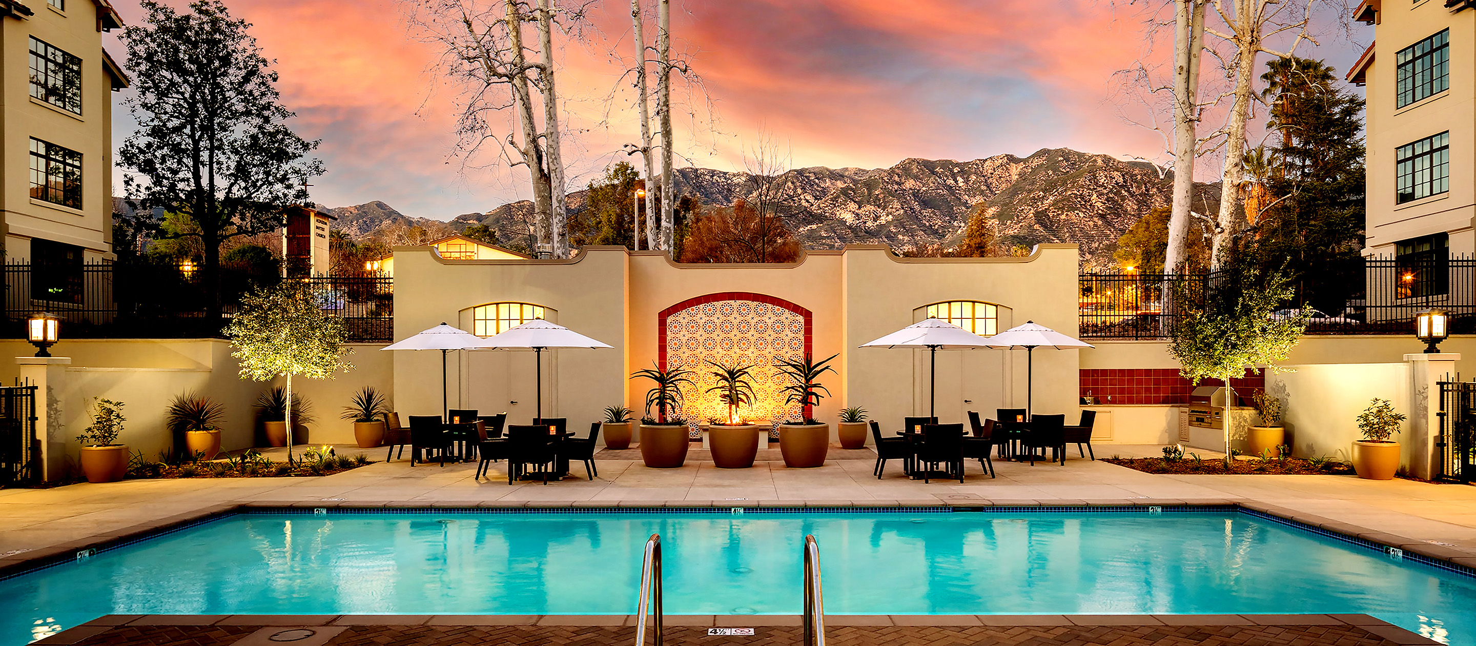 A swimming pool with lounge chairs and tables under white umbrellas, surrounded by potted plants. Beyond the pool, a decorative wall and mountain range are visible at sunset with a colorful sky.