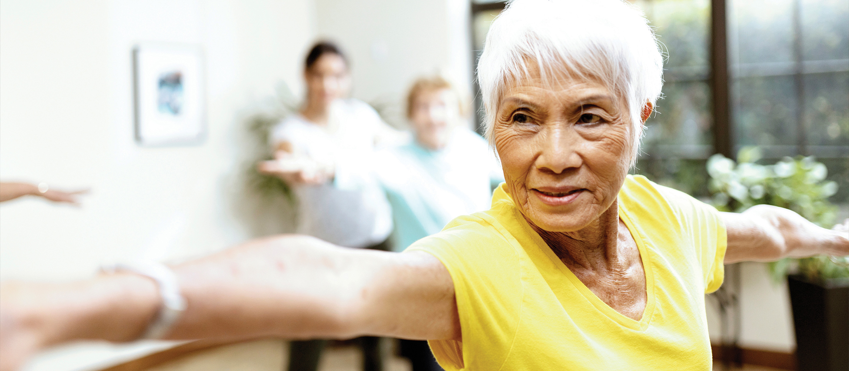 Older woman with short white hair, wearing a yellow shirt, stretches her arms out in a yoga pose indoors, with blurred people and plants in the background.