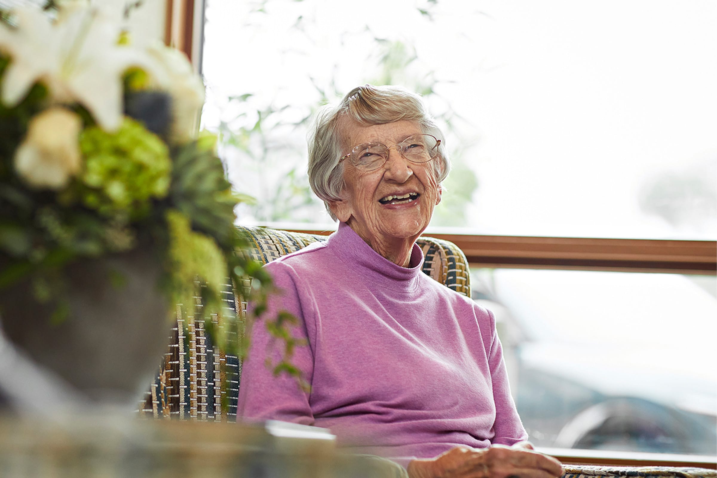 An elderly woman with short gray hair and glasses sits in a wicker chair, smiling warmly. She wears a light purple turtleneck sweater. Soft natural light comes through a window behind her, and a vase of flowers is in the foreground.