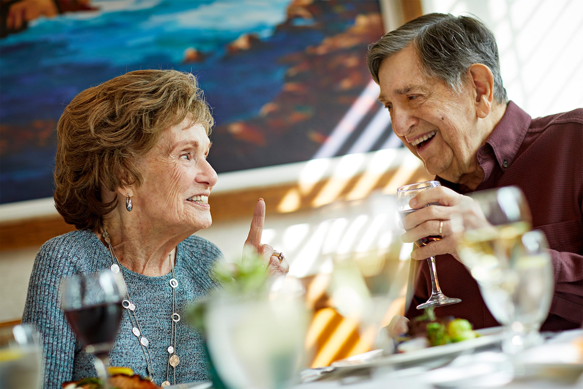 An elderly woman and man sit at a table in a restaurant, smiling and enjoying conversation. The woman gestures with her hand while the man holds up a glass. Food and drinks are on the table, and a painting is in the background.