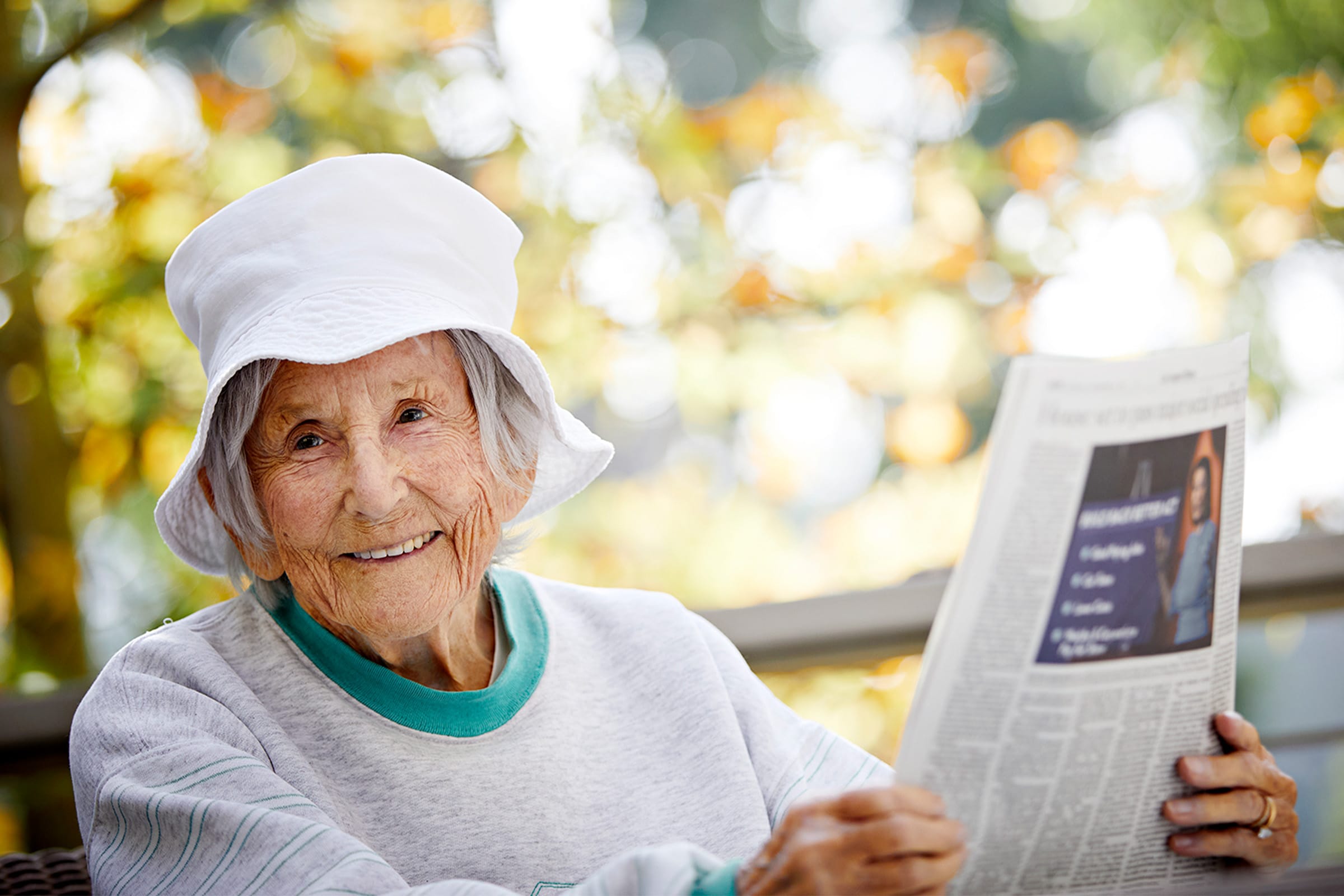 An elderly woman wearing a white sunhat and light sweater smiles while sitting outdoors, holding a newspaper. The background shows soft sunlight and blurred green foliage.