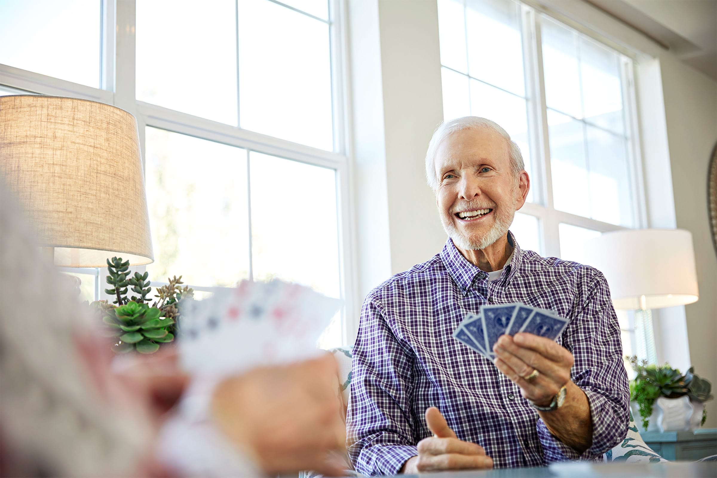 An older man with white hair, wearing a checkered shirt, smiles while holding playing cards at a table. Sunlight streams through large windows behind him, and another person with cards is partially visible in the foreground.