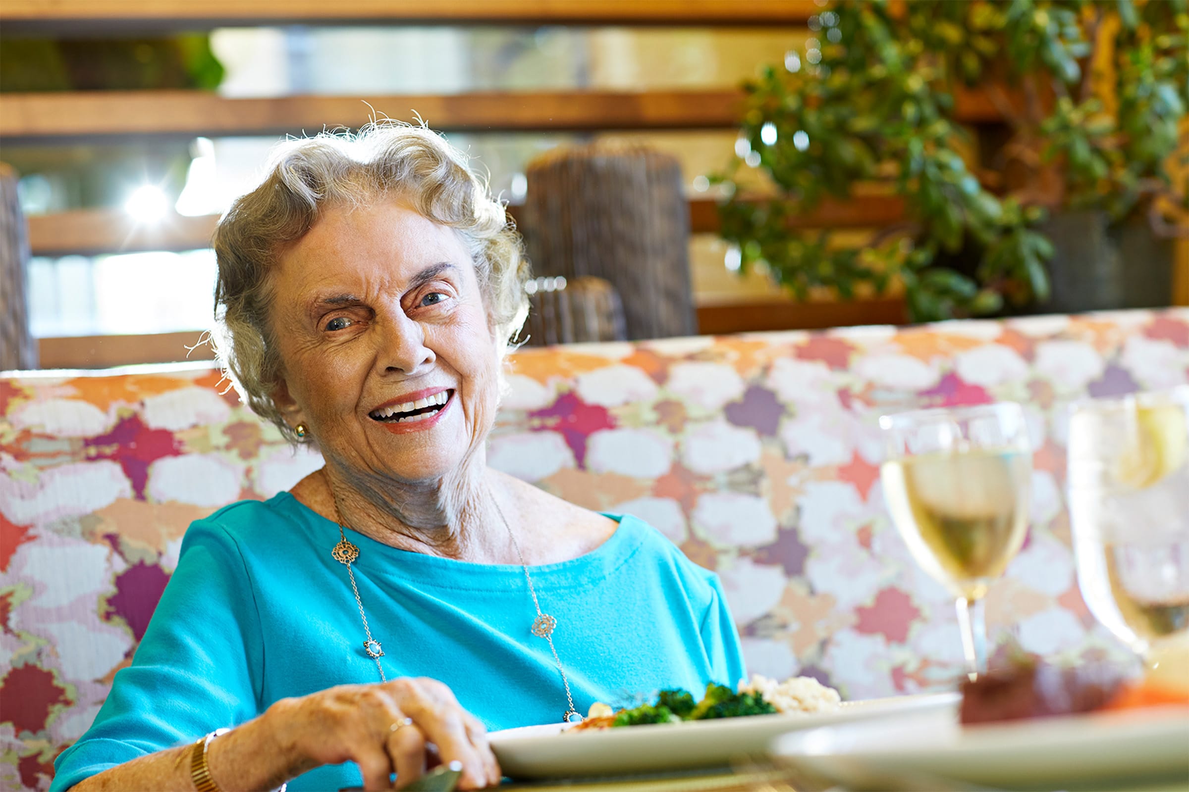 An elderly woman with short gray hair, wearing a turquoise top, smiles while sitting at a table with food and two glasses of white wine in a brightly lit, colorful restaurant.