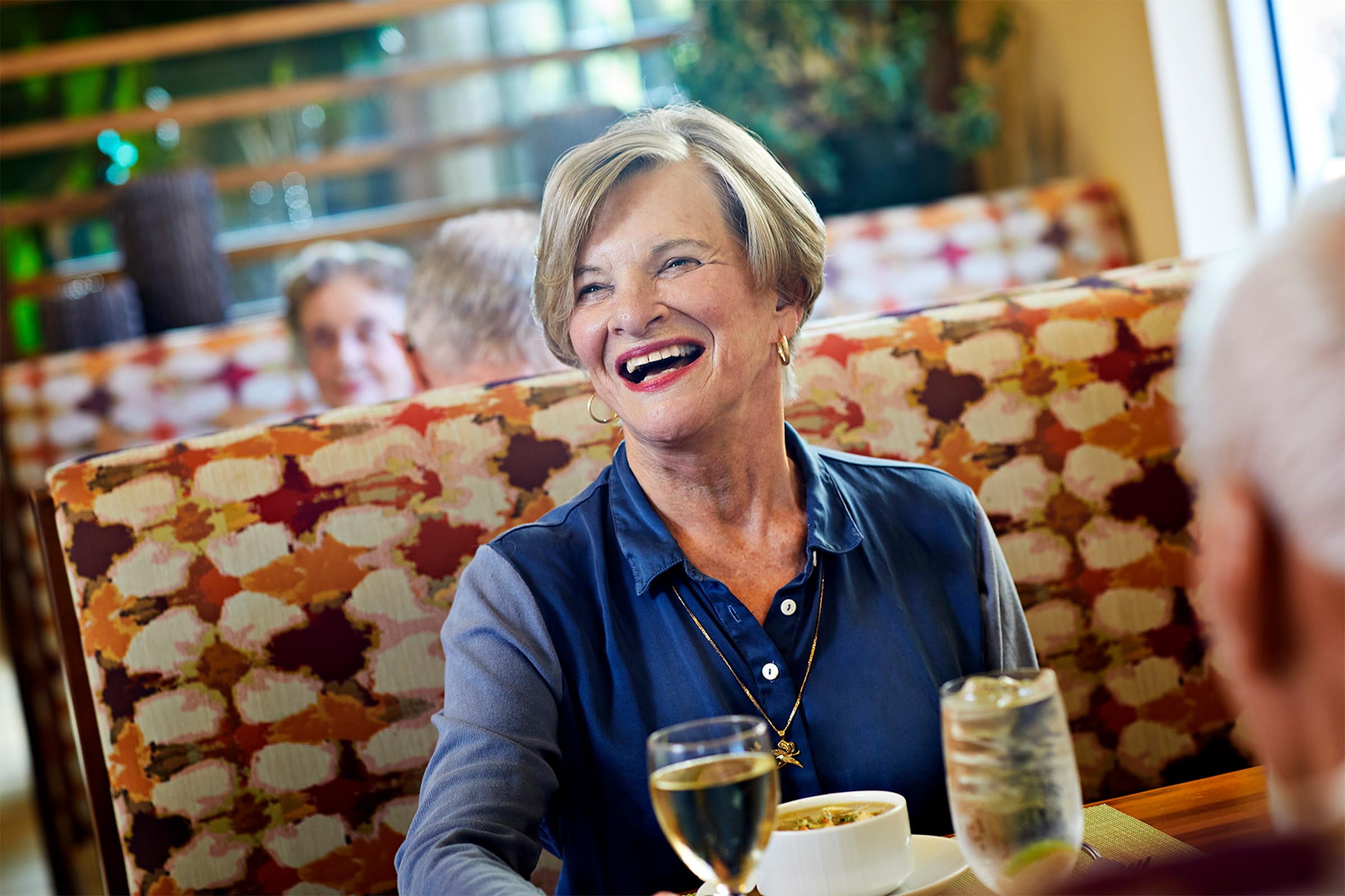 An older woman with short blond hair smiles while sitting at a restaurant booth. She is wearing a blue shirt and has a bowl of food, a glass of white wine, and a glass of water in front of her.