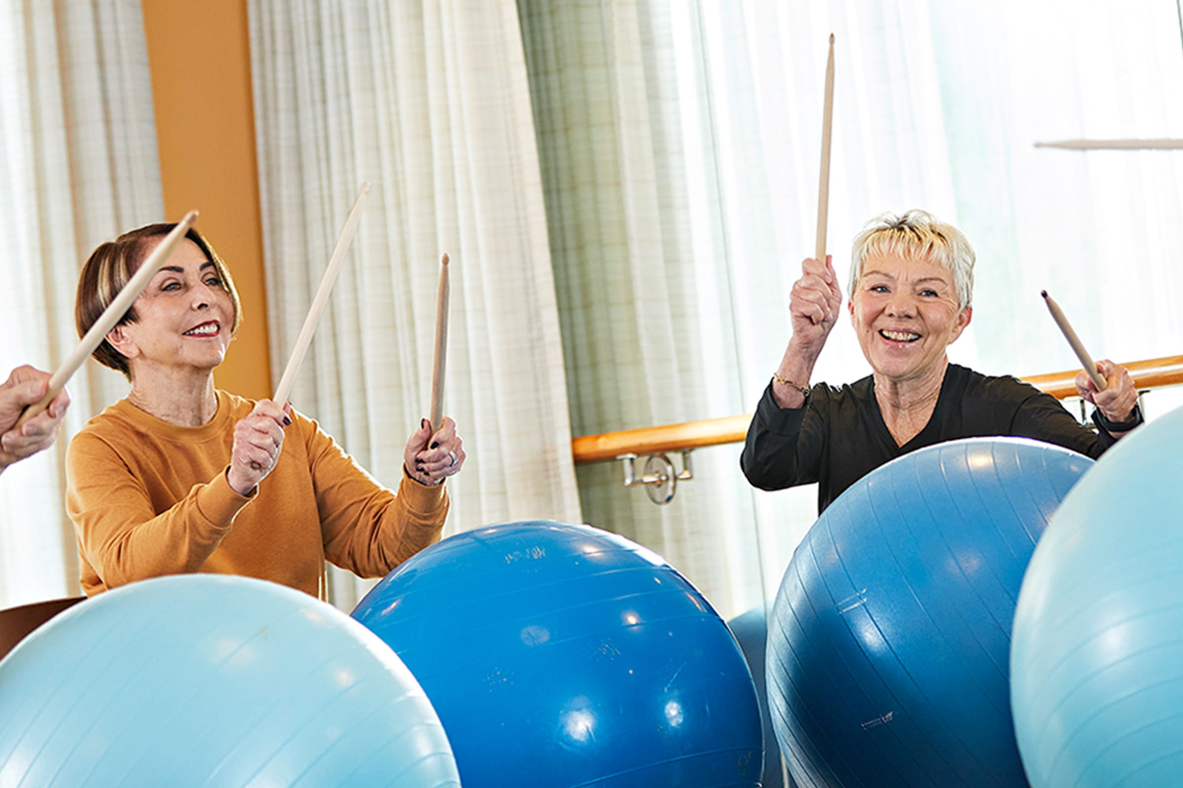 Two older women smile while participating in a fitness class, each hitting large blue exercise balls with drumsticks. They appear energetic and happy, with light streaming through curtains in the background.