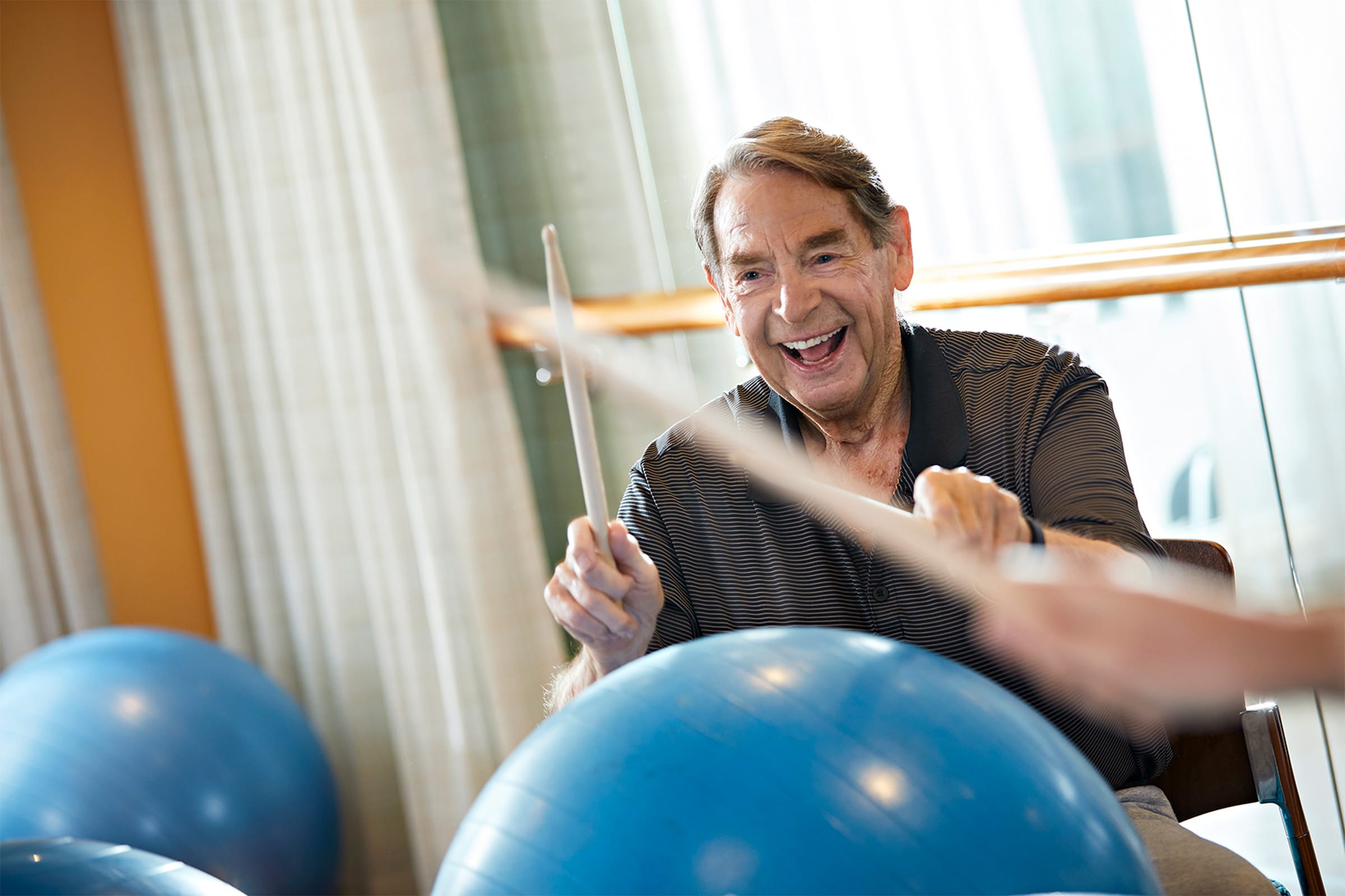 An older man smiles while using drumsticks to play on a large blue exercise ball in a bright room, engaging in a fun and active exercise.