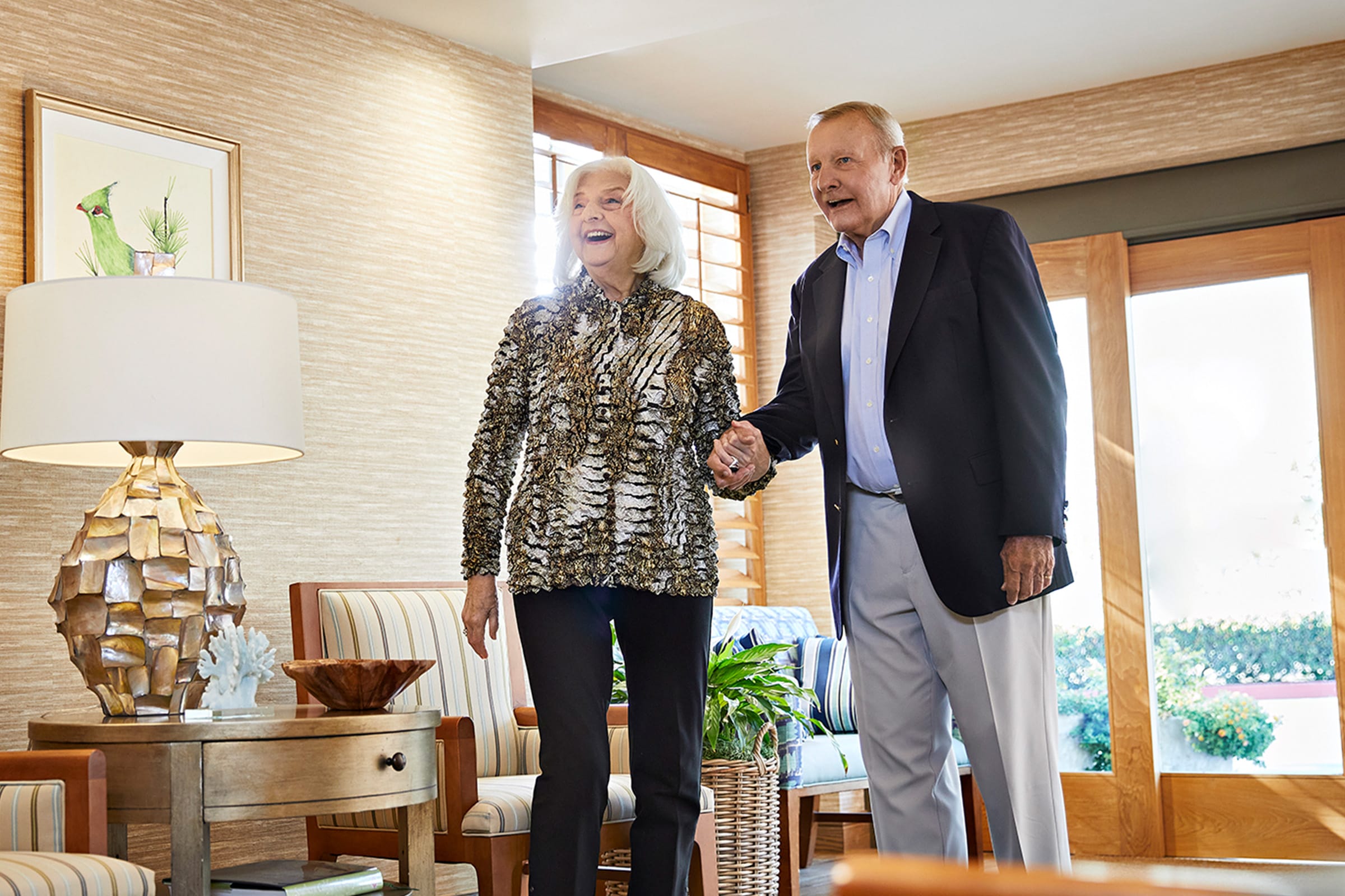 An older couple smiles while standing and holding hands in a bright, cozy living room with wood furniture, striped chairs, plants, and large windows in the background.
