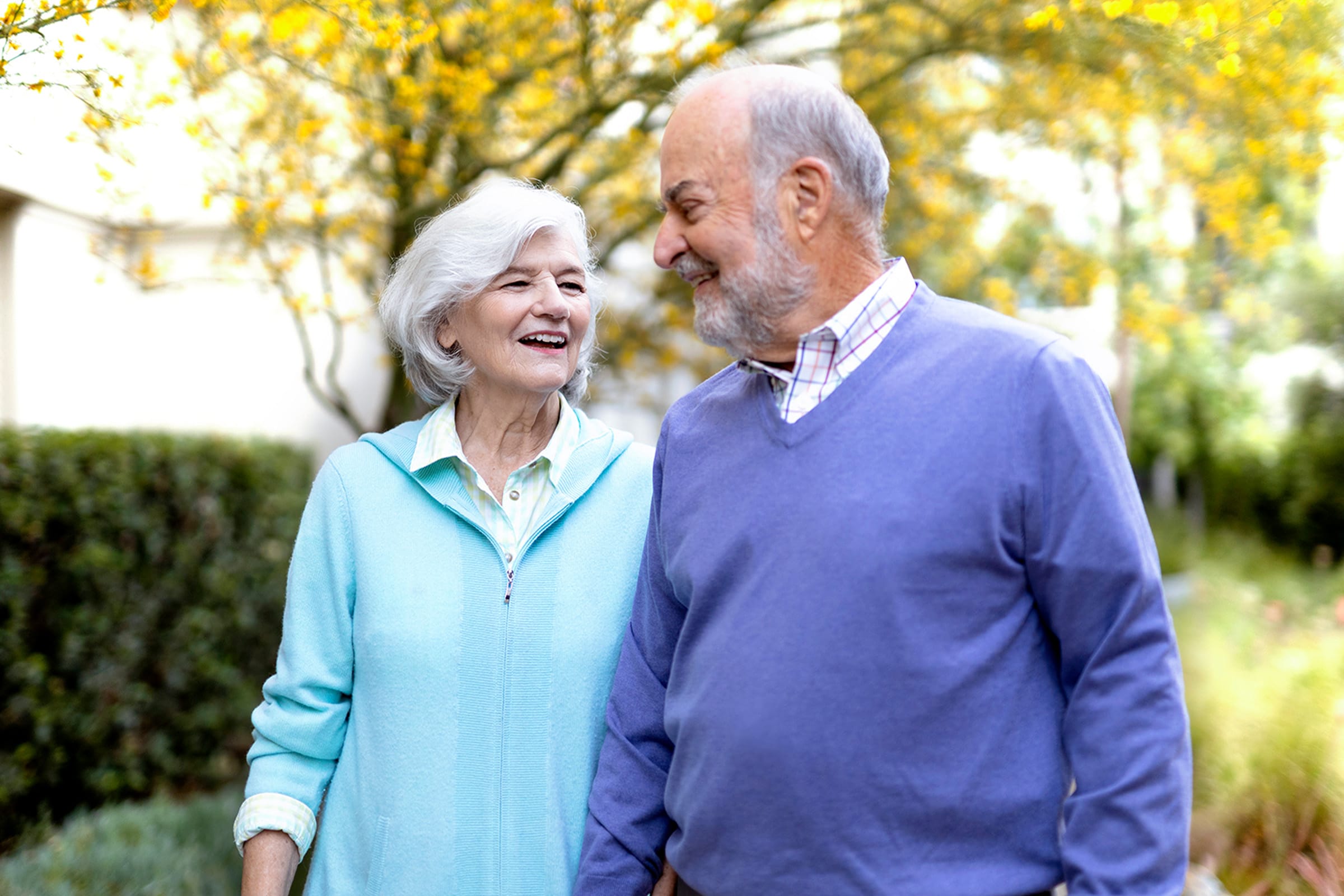 An older woman and man smile warmly at each other while walking outdoors. They are wearing light sweaters and are surrounded by greenery and yellow-blossomed trees, enjoying a pleasant day together.