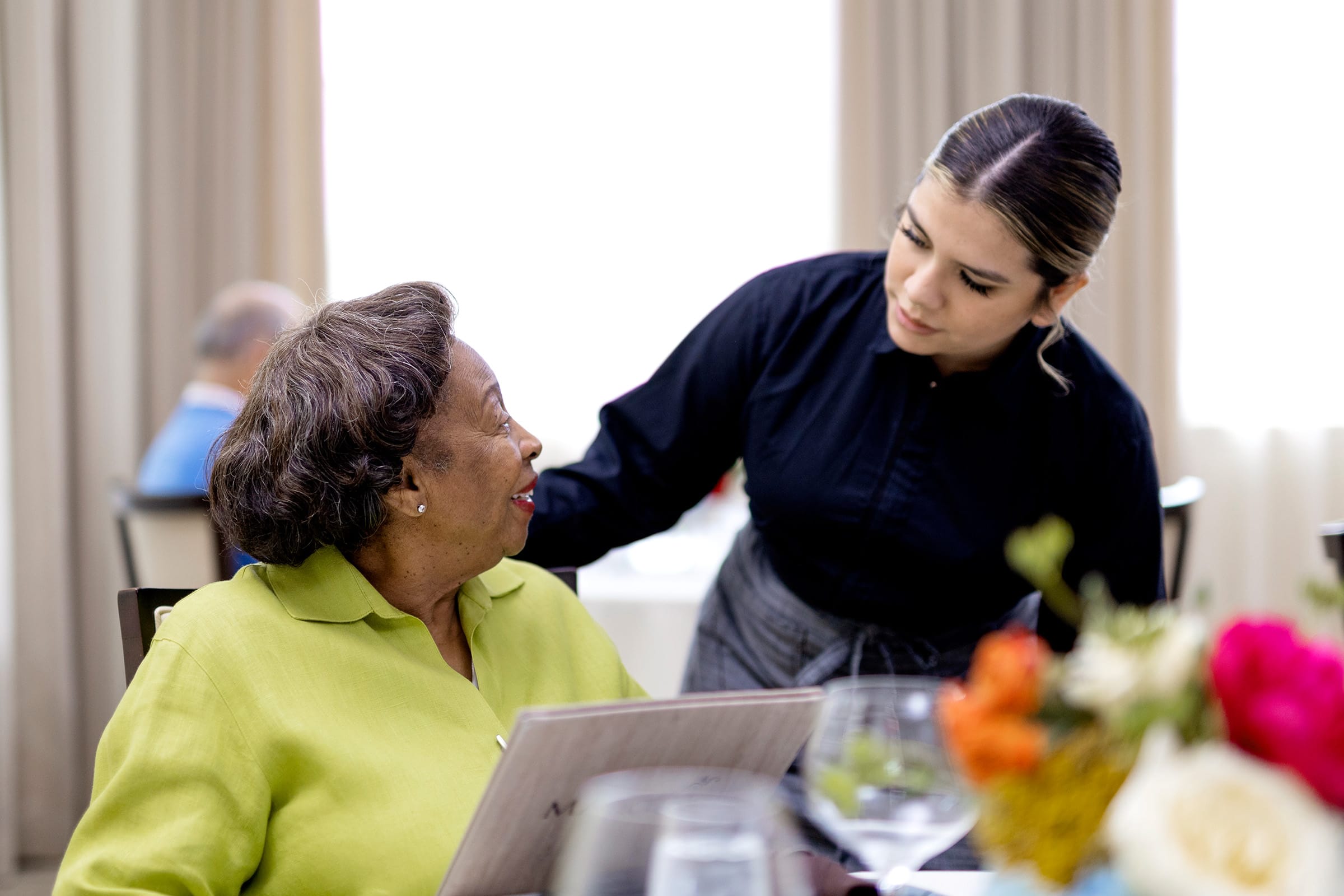 An elderly woman in a green blouse holds a menu and smiles while talking to a waitress in a black uniform at a restaurant table, with flowers and glasses visible in the foreground.