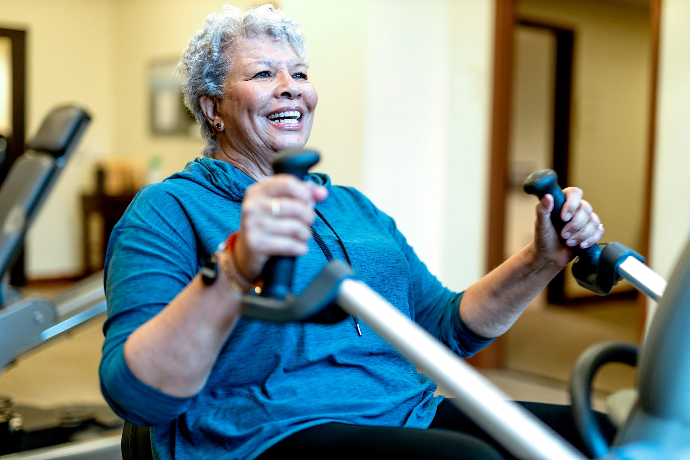 An older woman with short gray hair smiles while exercising on a rowing machine in a gym, wearing a blue hoodie and appearing energetic and happy.