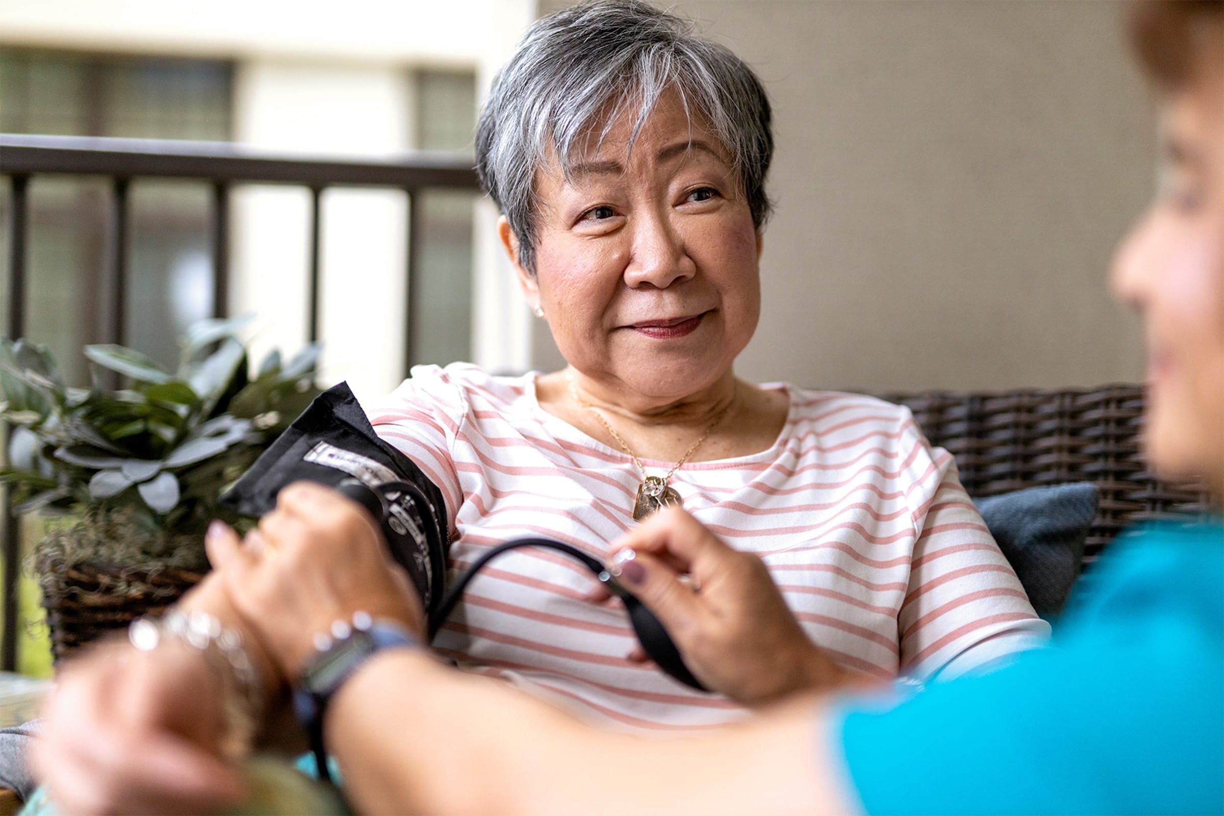 An older woman with short gray hair sits on a couch, smiling, while another person checks her blood pressure with a cuff on her arm. A potted plant is visible in the background.