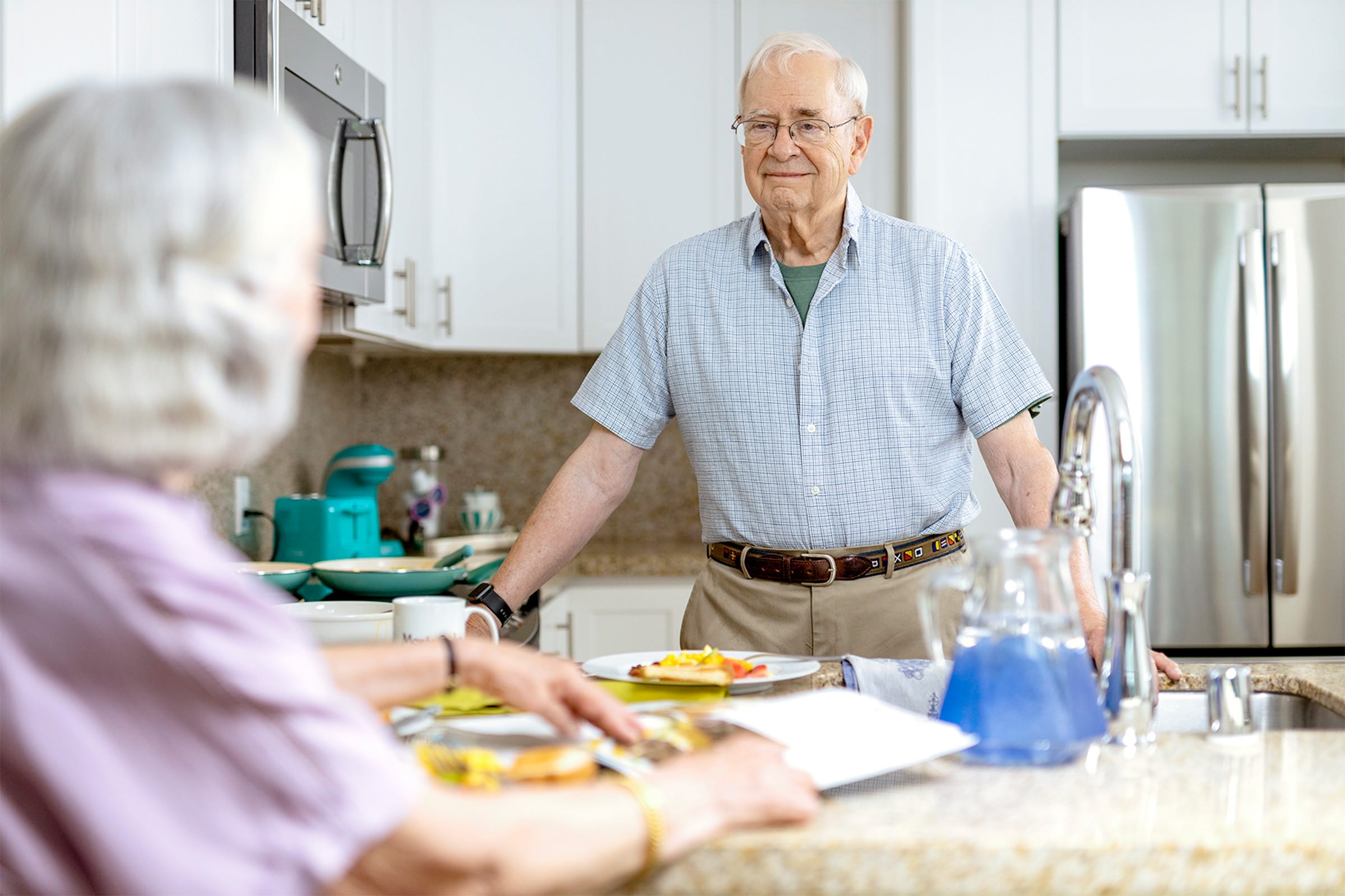 An elderly man stands in a bright kitchen, smiling at an elderly woman seated at the table. Breakfast is served on the counter, and a glass pitcher with blue liquid sits by the sink.