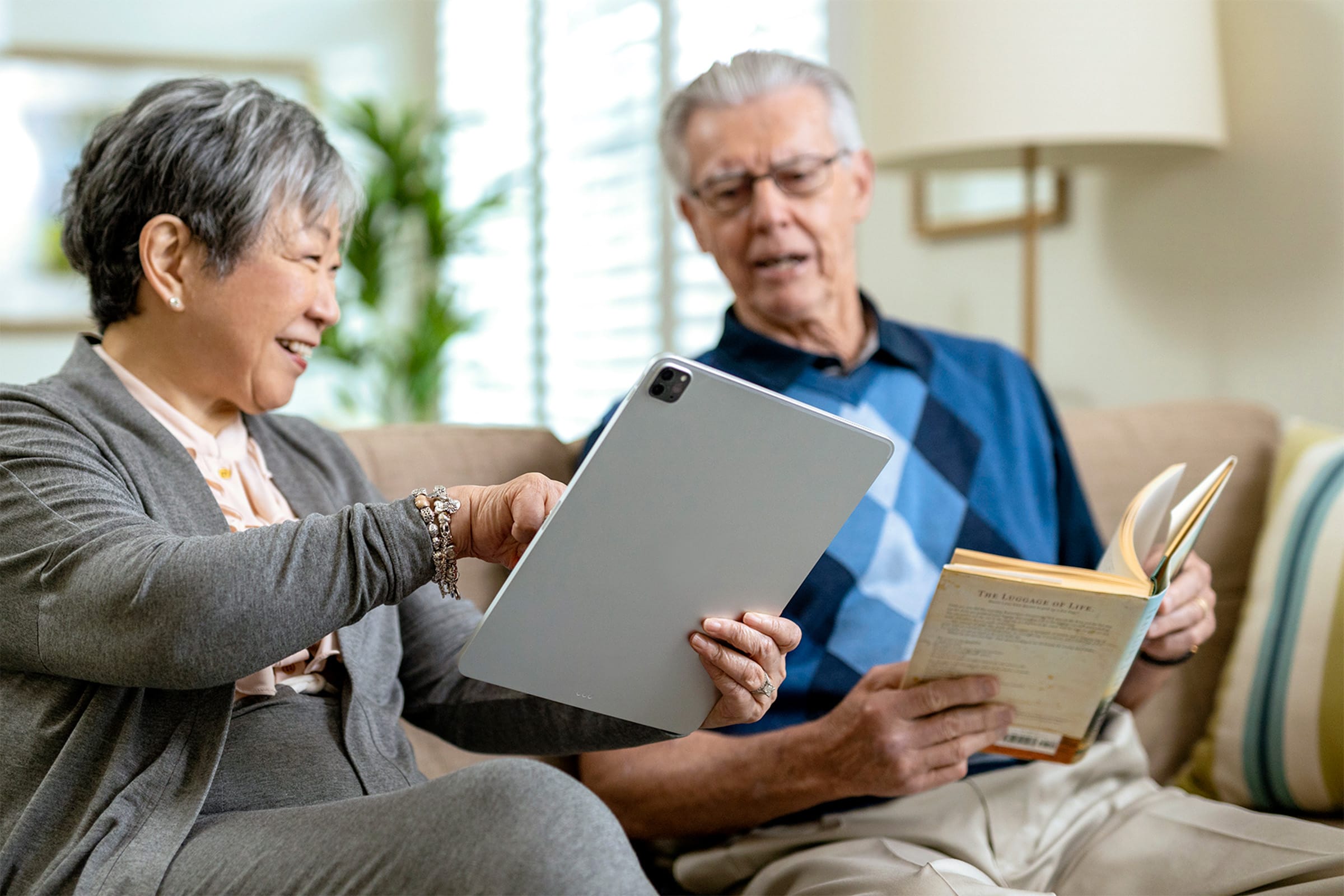 An elderly woman smiles while showing something on a tablet to an elderly man sitting beside her. He is holding an open book. They are both seated on a couch in a brightly lit living room.