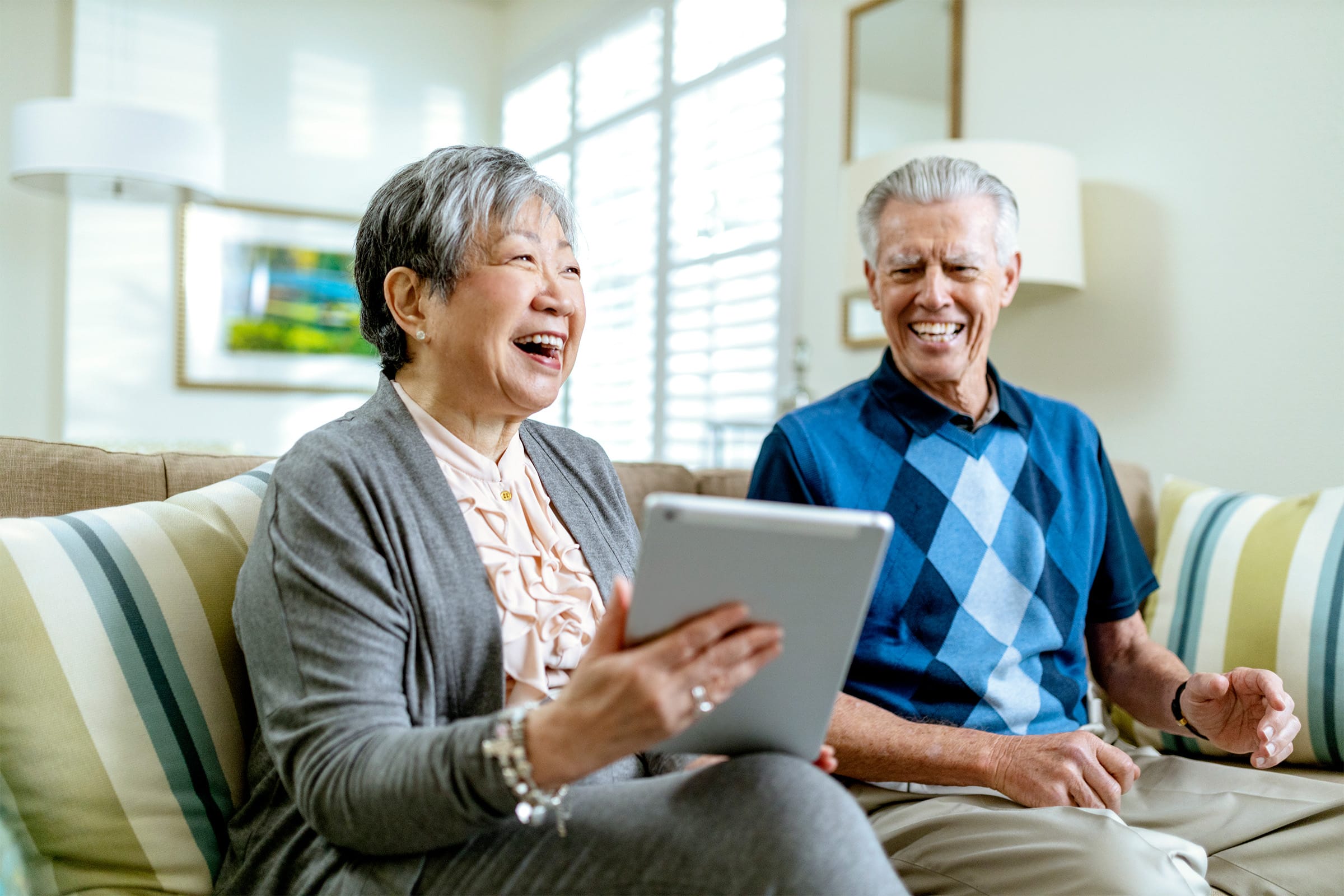 An older woman and man sit on a couch, smiling and laughing together. The woman holds a tablet, and both appear to be enjoying their time in a bright, cozy living room.