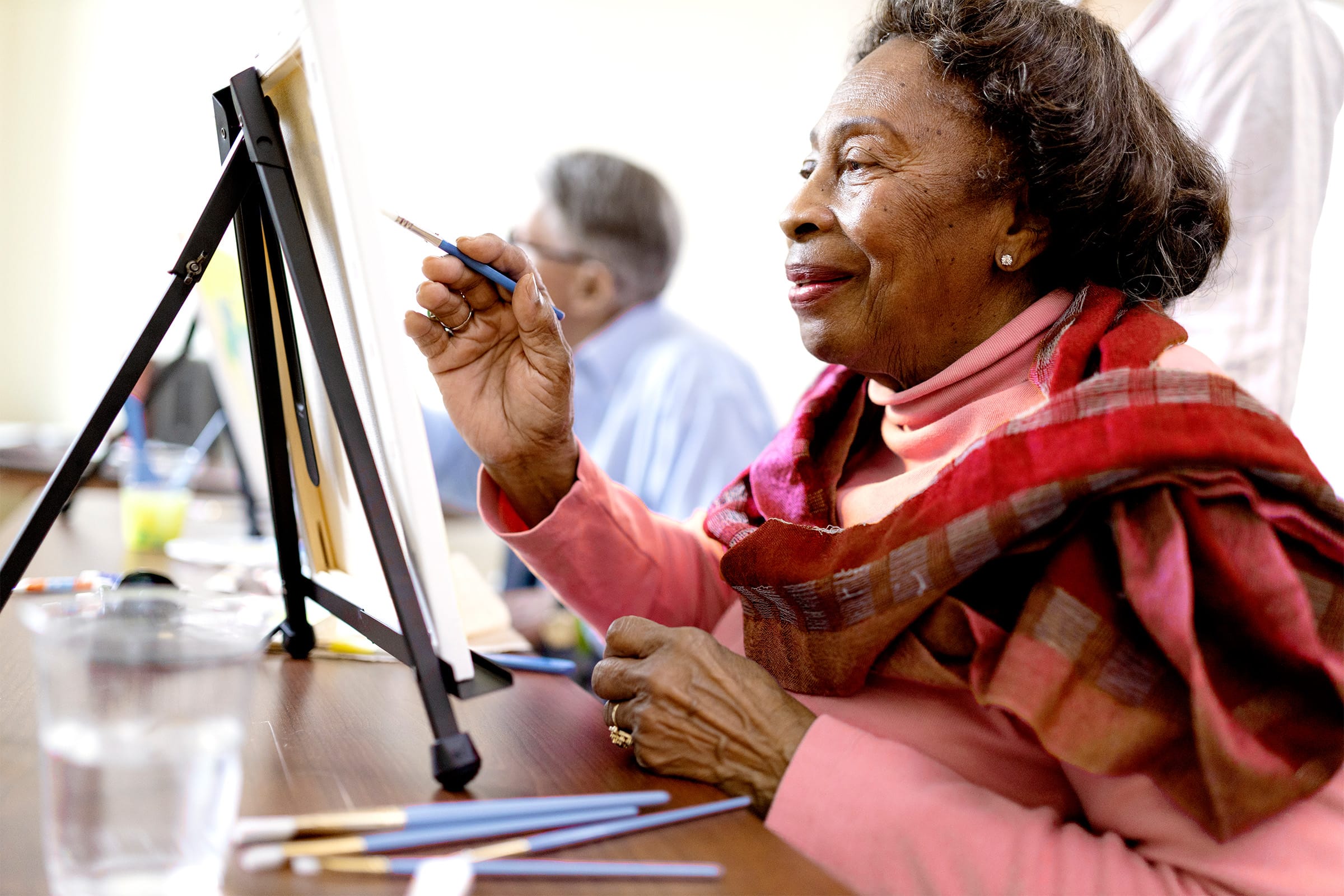 An older woman wearing a pink turtleneck and shawl smiles while painting at an easel, with brushes and a glass of water on the table. Another person is blurred in the background.