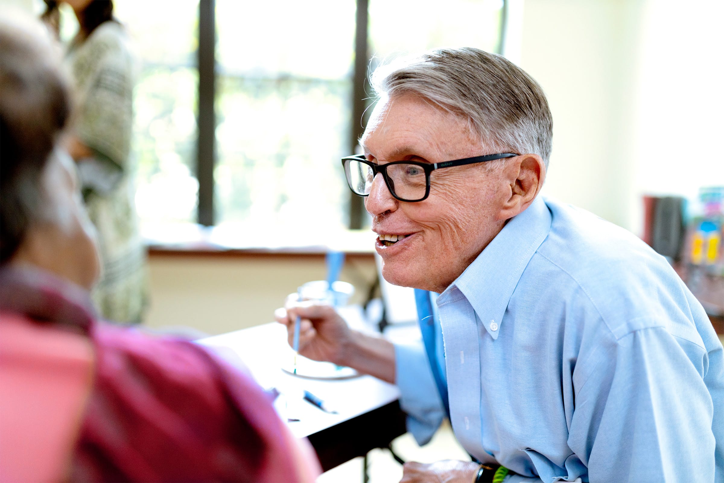 An older man with gray hair and glasses smiles and converses with another person indoors. He is wearing a light blue shirt and appears engaged and friendly. The background is softly lit with windows.