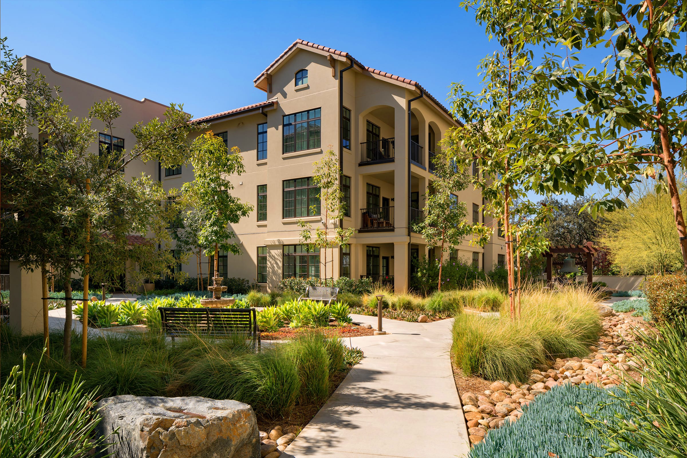 A modern, beige three-story apartment building with balconies, surrounded by landscaped greenery, trees, rocks, and a winding concrete pathway under a clear blue sky.