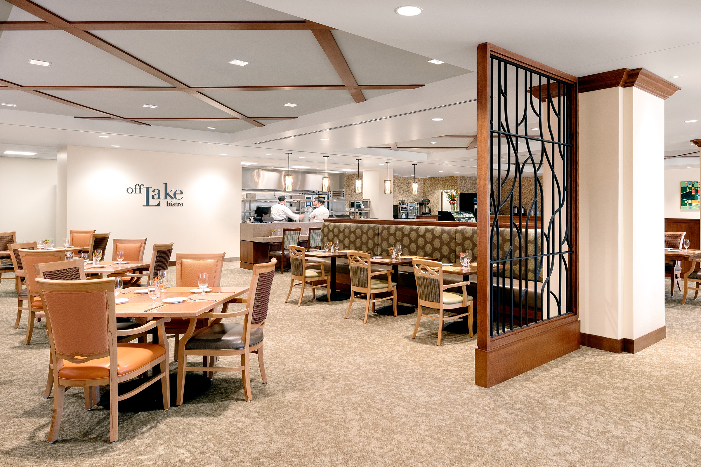 A modern, well-lit restaurant with tan and wooden decor, neatly set tables and chairs, cushioned booths, and an open kitchen where chefs are preparing food. A decorative metal divider separates dining areas.