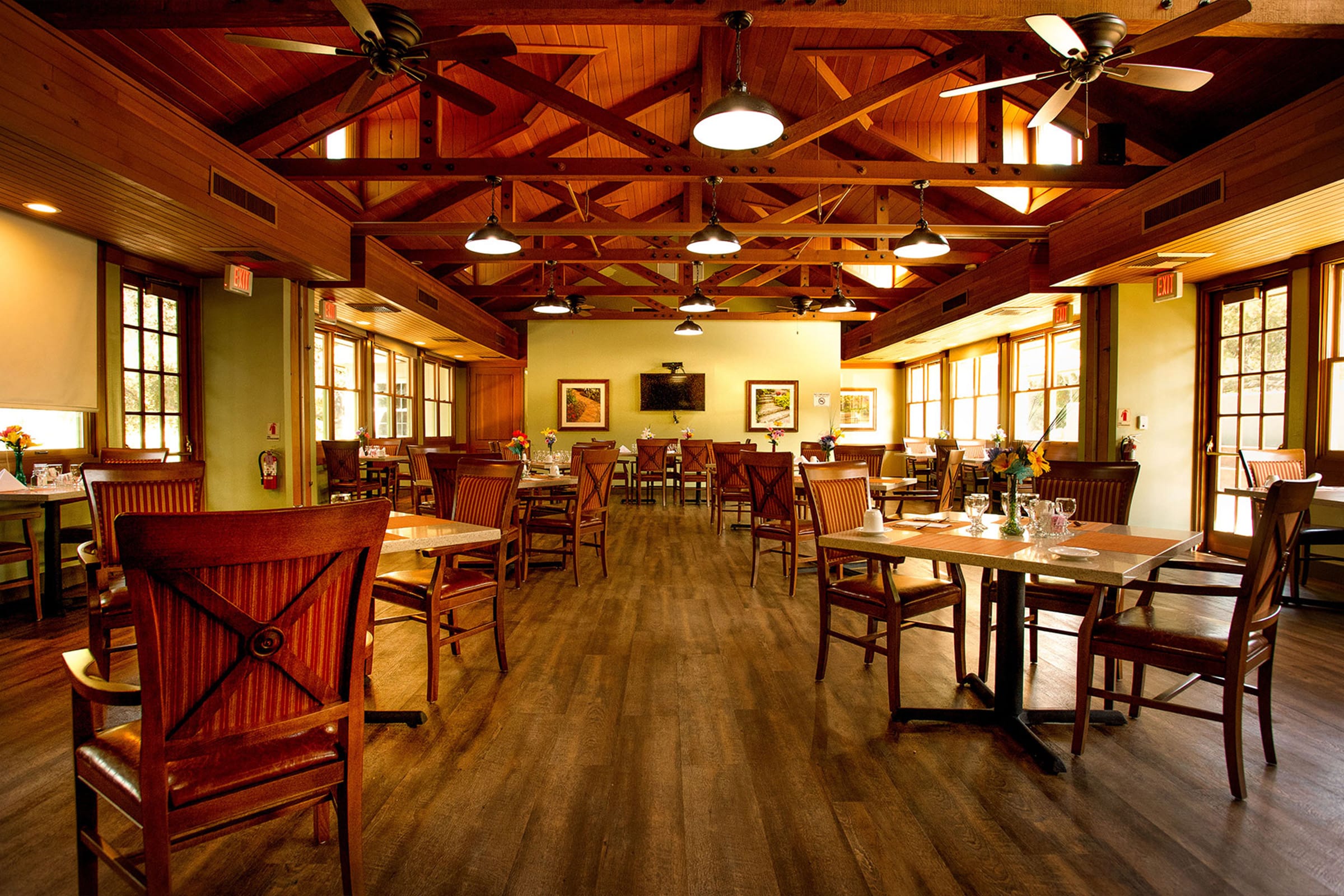 A spacious, warmly lit restaurant dining area with wooden floors, high wooden ceilings, ceiling fans, and large windows. Wooden tables and chairs are neatly arranged, set with glasses and cutlery.
