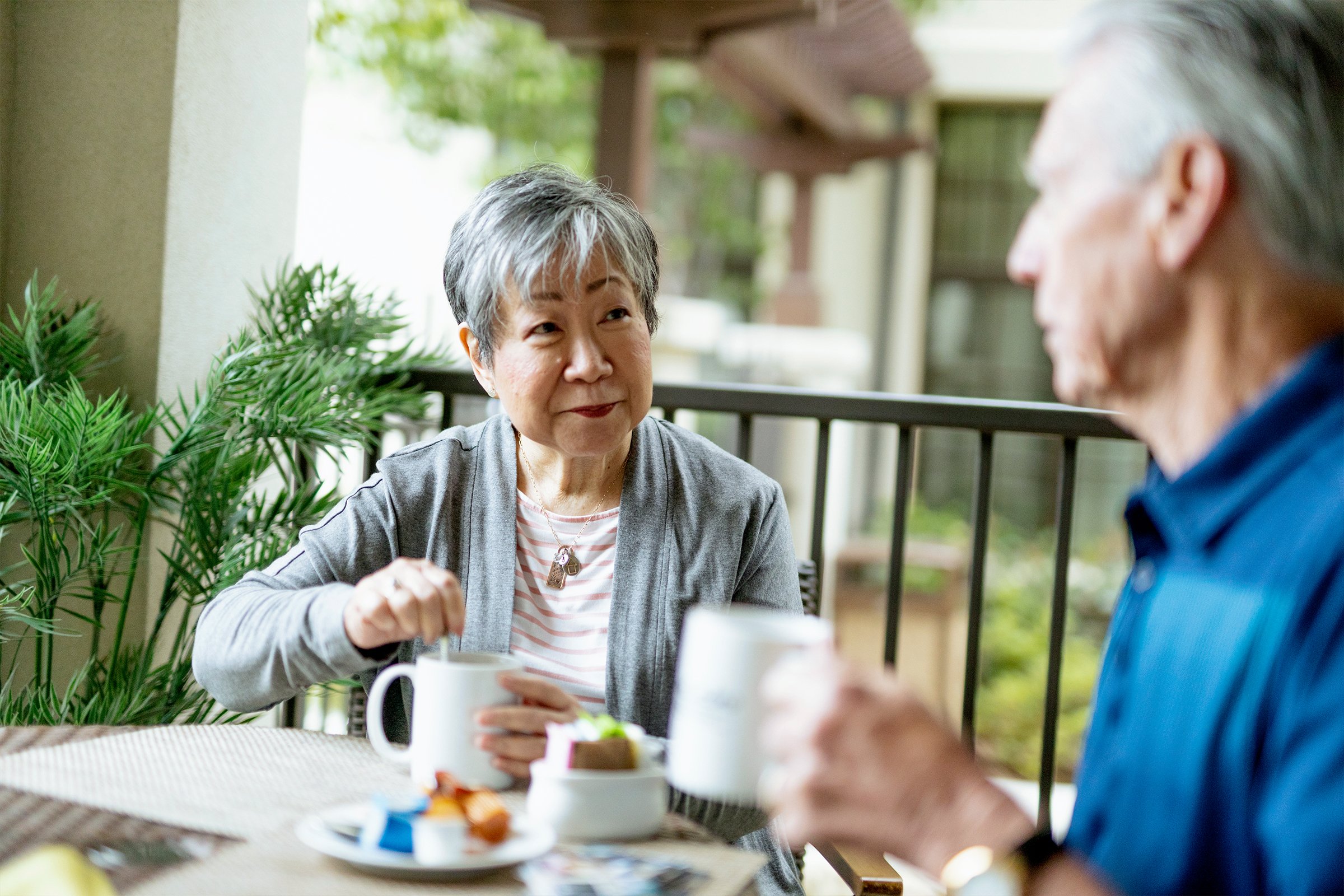 An older woman stirs a drink while sitting at an outdoor table with an older man. Both hold white mugs and appear to be enjoying a relaxed conversation. There are snacks and greenery in the background.