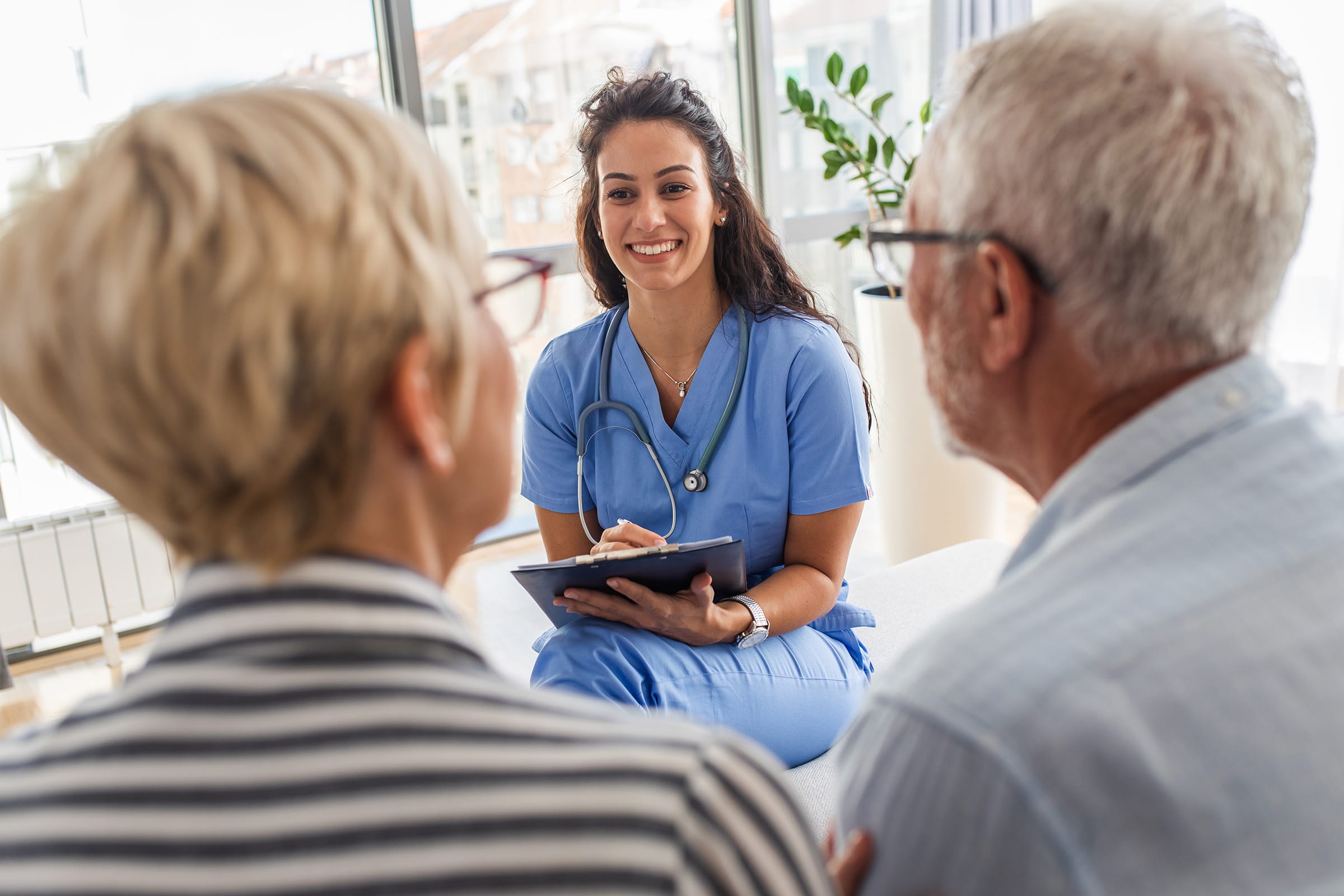 A smiling healthcare worker in blue scrubs talks with an older couple, holding a clipboard and pen, in a bright room with large windows and a potted plant in the background.