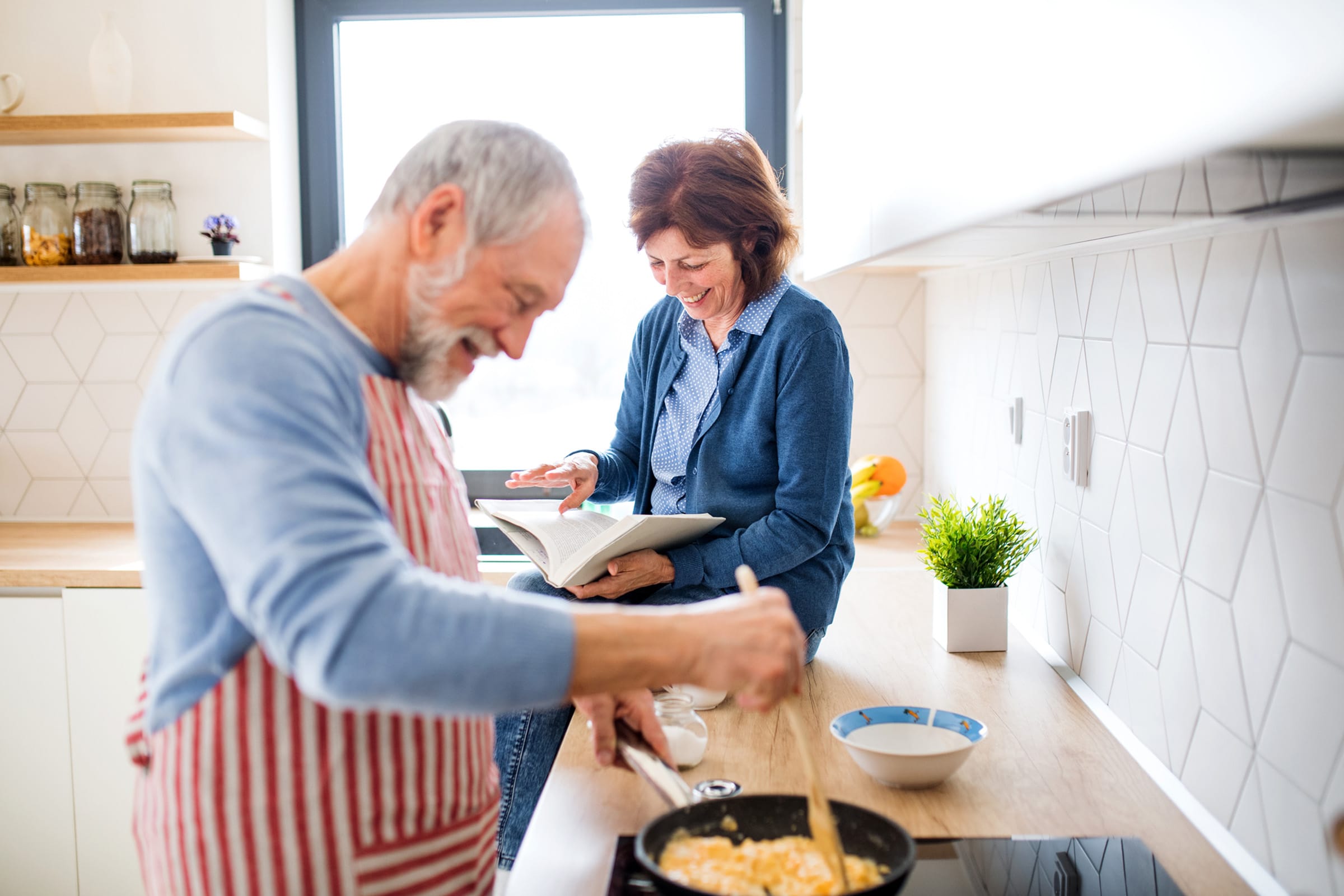 An older man in an apron cooks scrambled eggs in a pan while a woman smiles and reads a cookbook at the kitchen counter in a bright, modern kitchen.