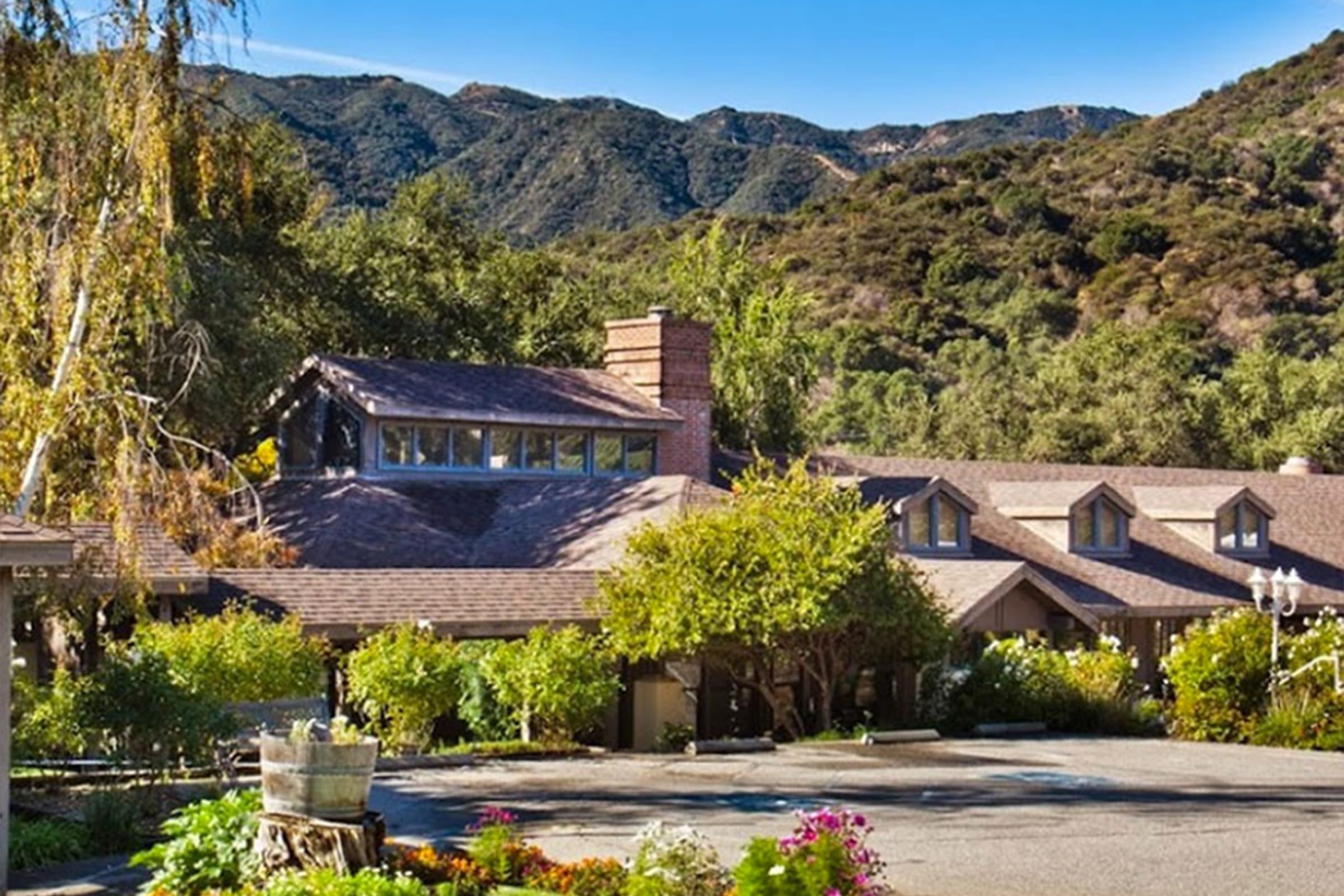 A rustic house with multiple gabled roofs and a large chimney sits surrounded by lush trees and gardens, with mountains and a clear blue sky in the background.