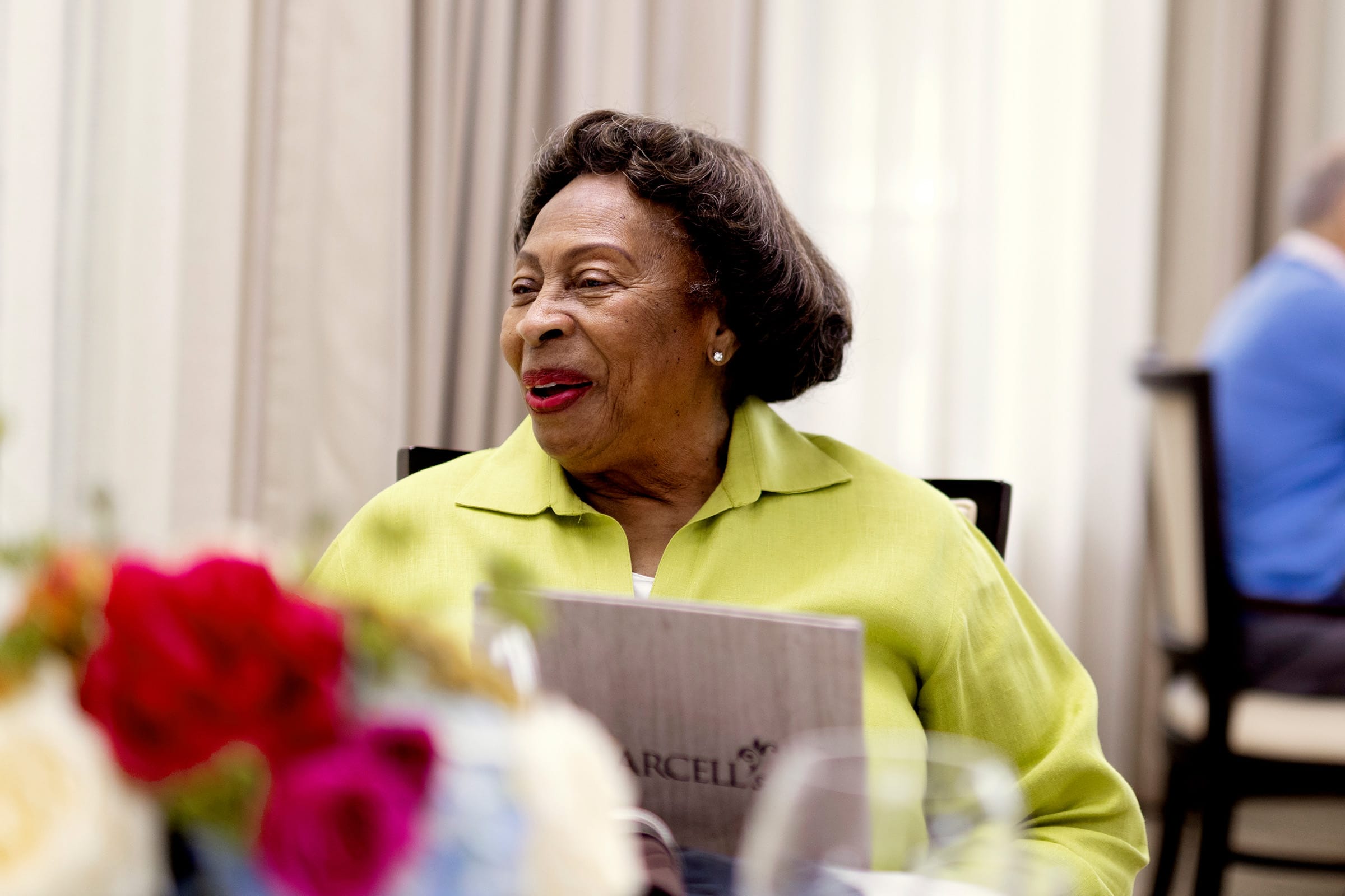 _photo-call-home An elderly woman in a light green shirt smiles while sitting at a table with a menu and colorful flowers in the foreground. The background is softly blurred.