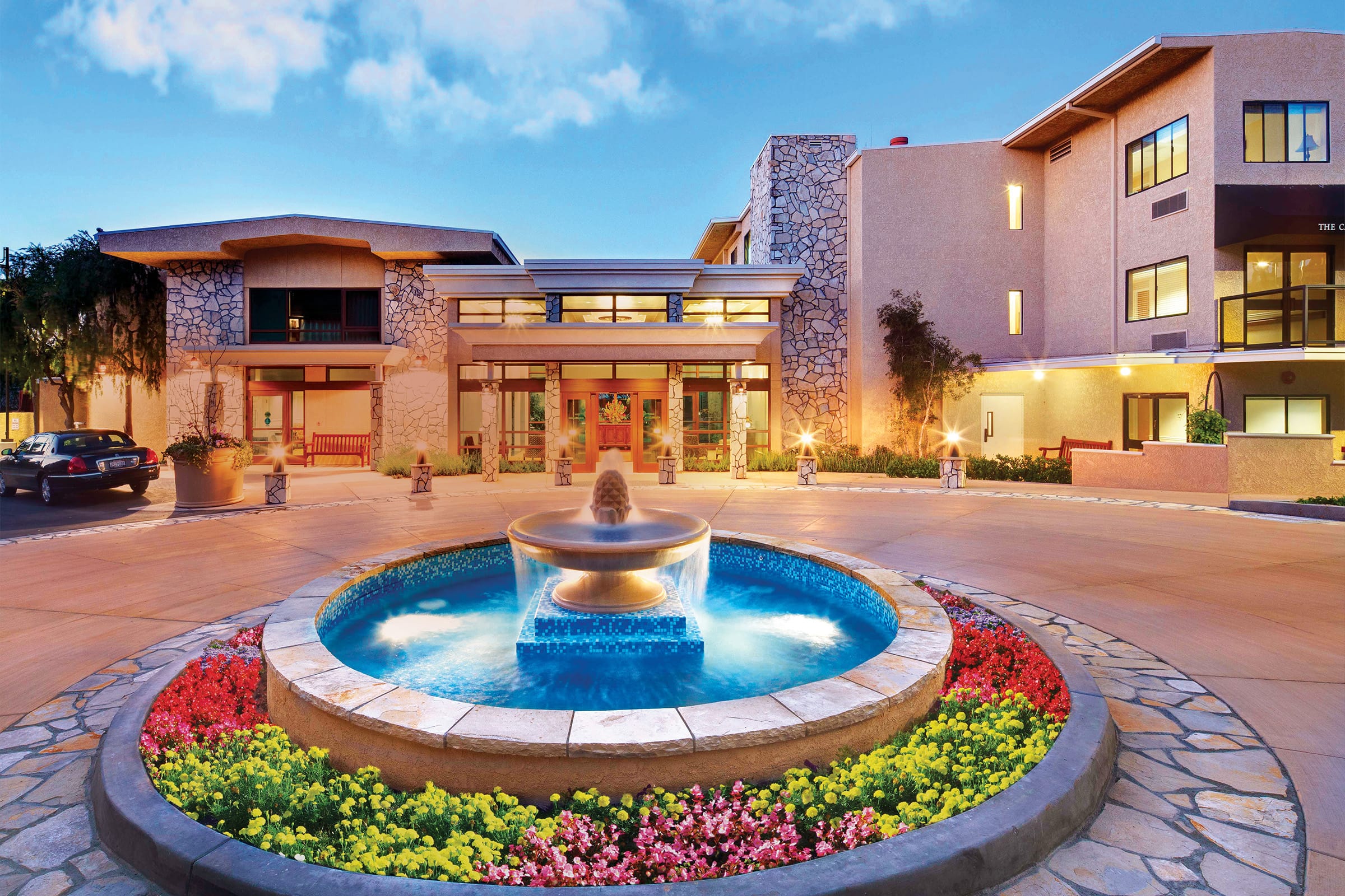 A modern building with large windows and stone accents features a circular fountain in the foreground, surrounded by colorful flowers and neatly landscaped greenery, under a blue sky at dusk.