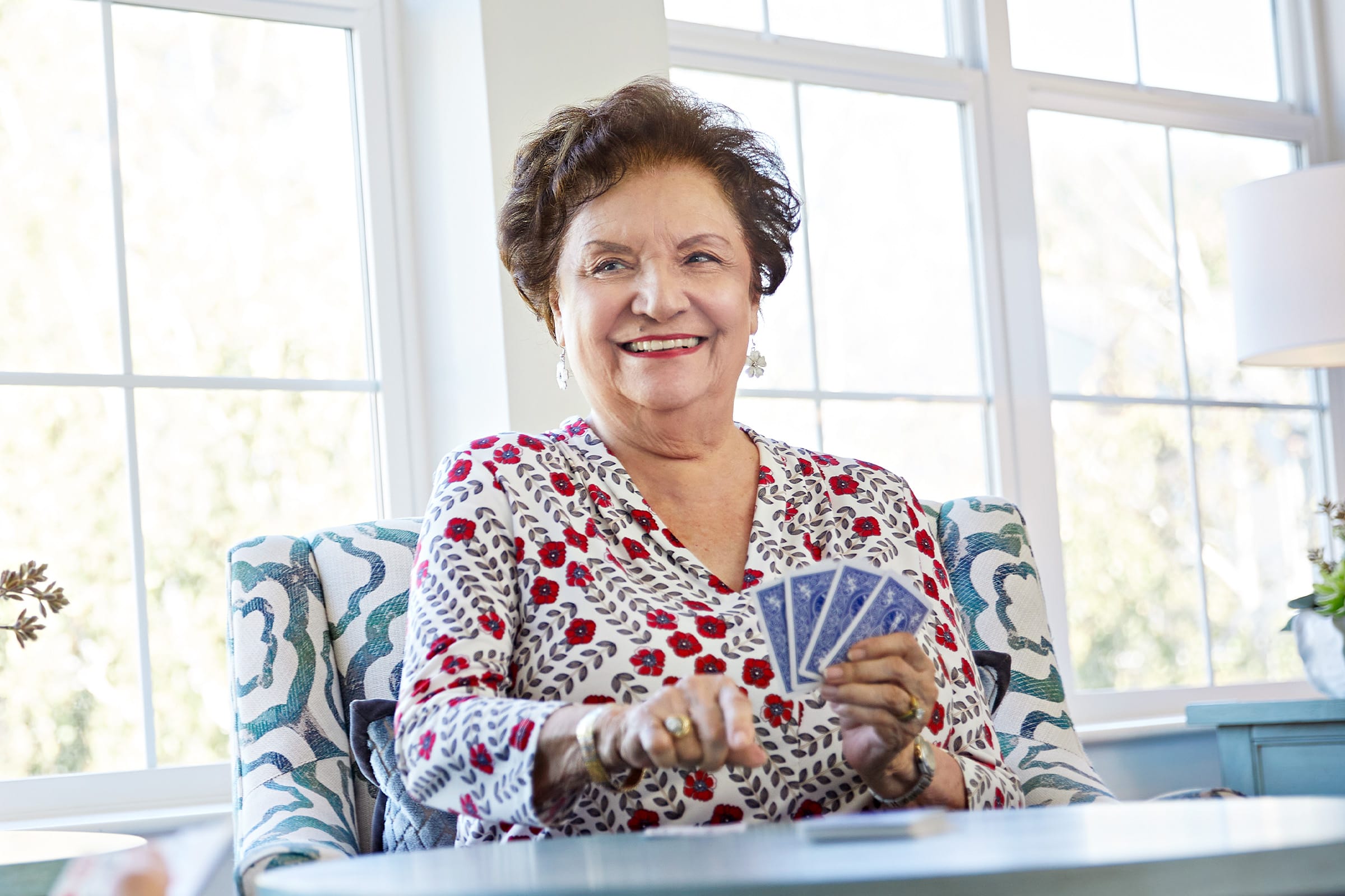 An older woman with short brown hair smiles while sitting at a table holding playing cards. She wears a patterned blouse and is seated in front of large windows with sunlight streaming in.