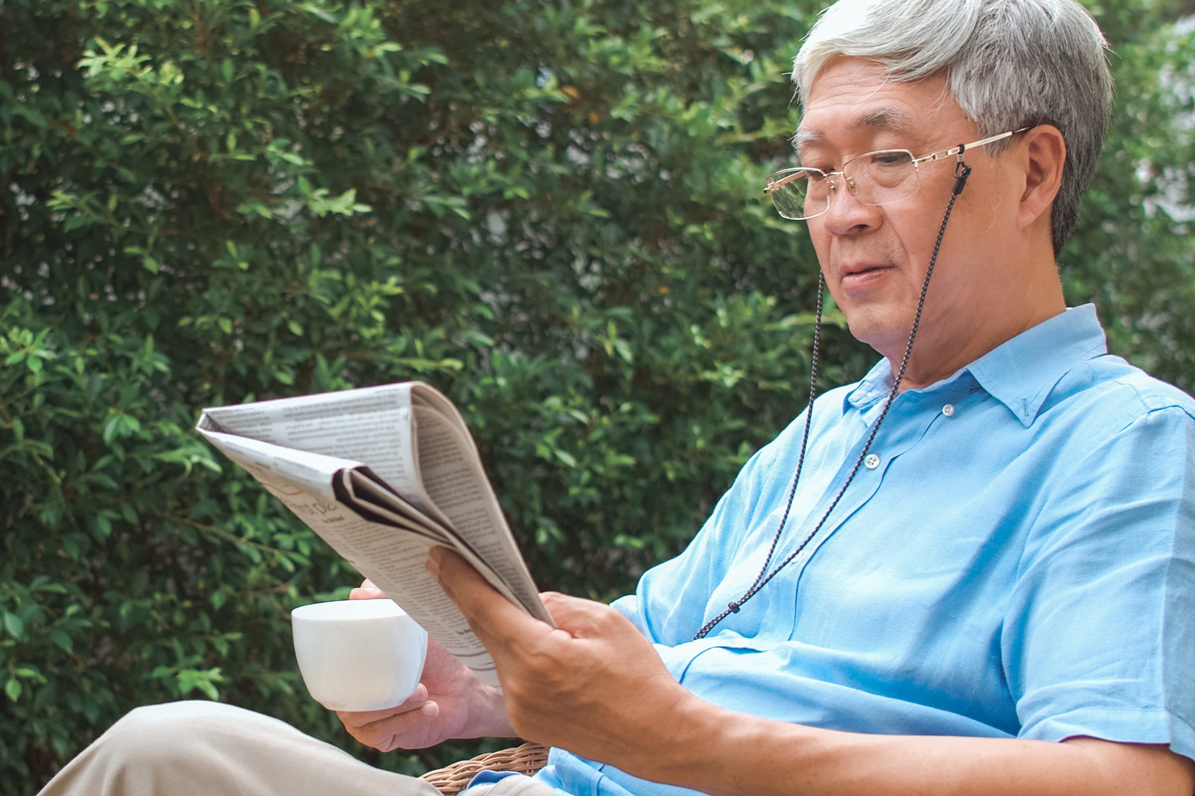 An older man with gray hair and glasses reads a newspaper and holds a white mug while sitting outdoors, surrounded by greenery. He is wearing a light blue shirt and appears relaxed.