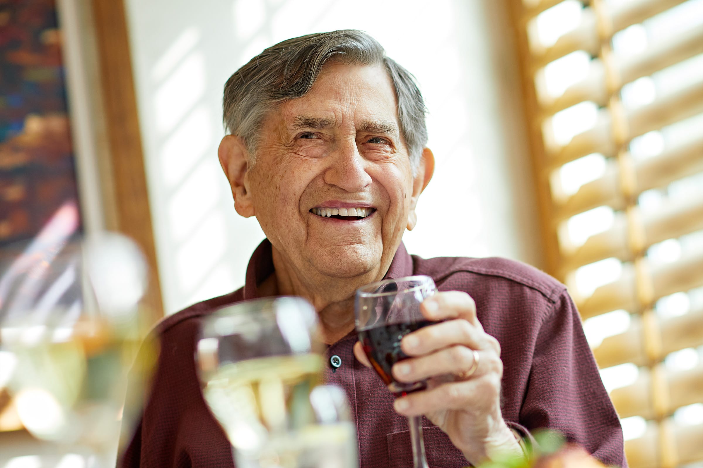 _photo-create An elderly man with gray hair smiles brightly while holding a glass of red wine. He is seated indoors at a table with blurred glasses and warm sunlight streaming through window blinds behind him.