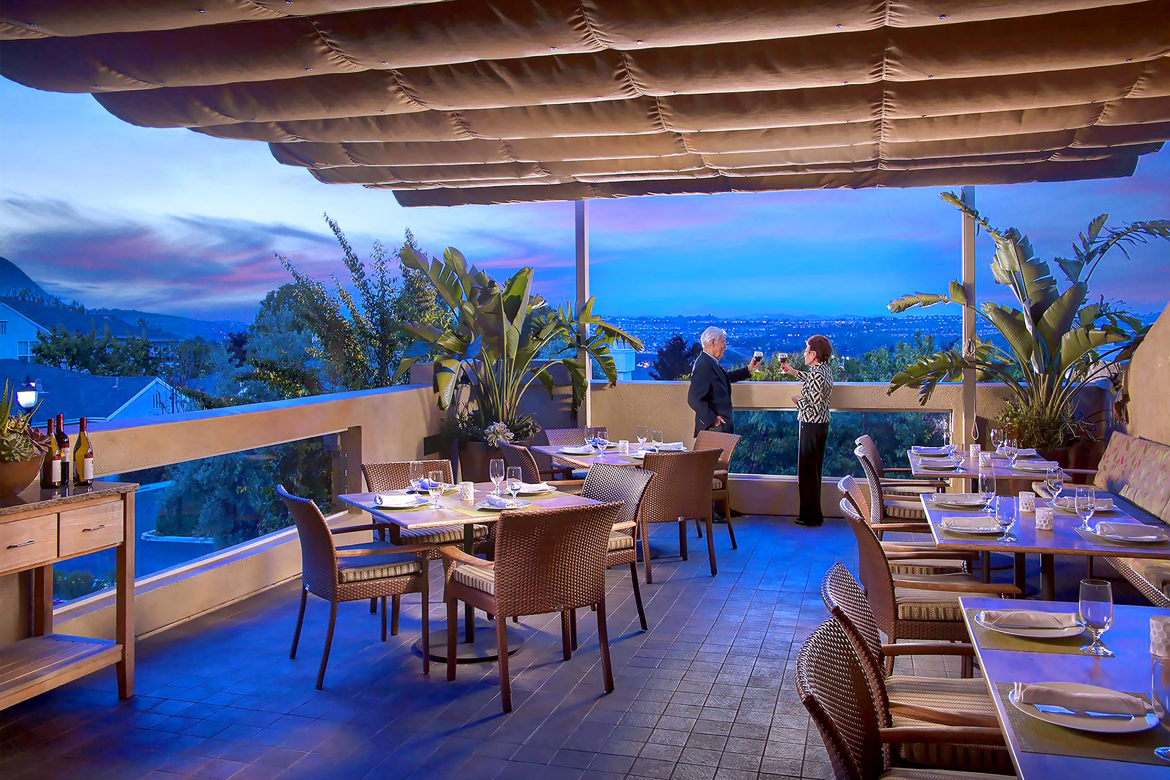 An elegant outdoor restaurant terrace at sunset, with tables set for dining, lush plants, and two people standing by the railing enjoying the scenic city view in the background.