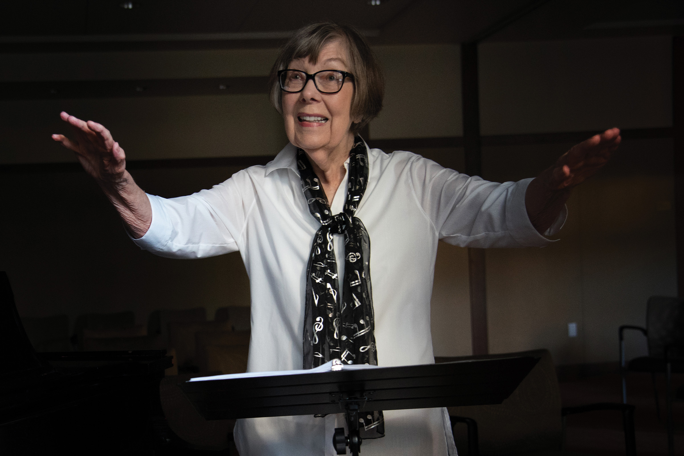 An older woman wearing glasses, a white shirt, and a black scarf stands in front of a music stand, smiling with her arms raised as if conducting or instructing. The background is dimly lit.