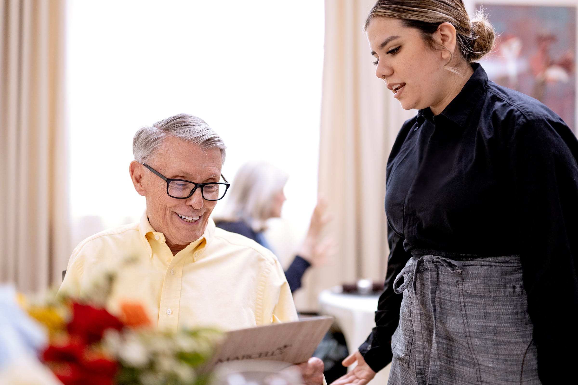 An older man wearing glasses smiles while looking at a menu, as a waitress in a black shirt and gray apron stands beside him in a bright, elegant restaurant. A bouquet of flowers is in the foreground.