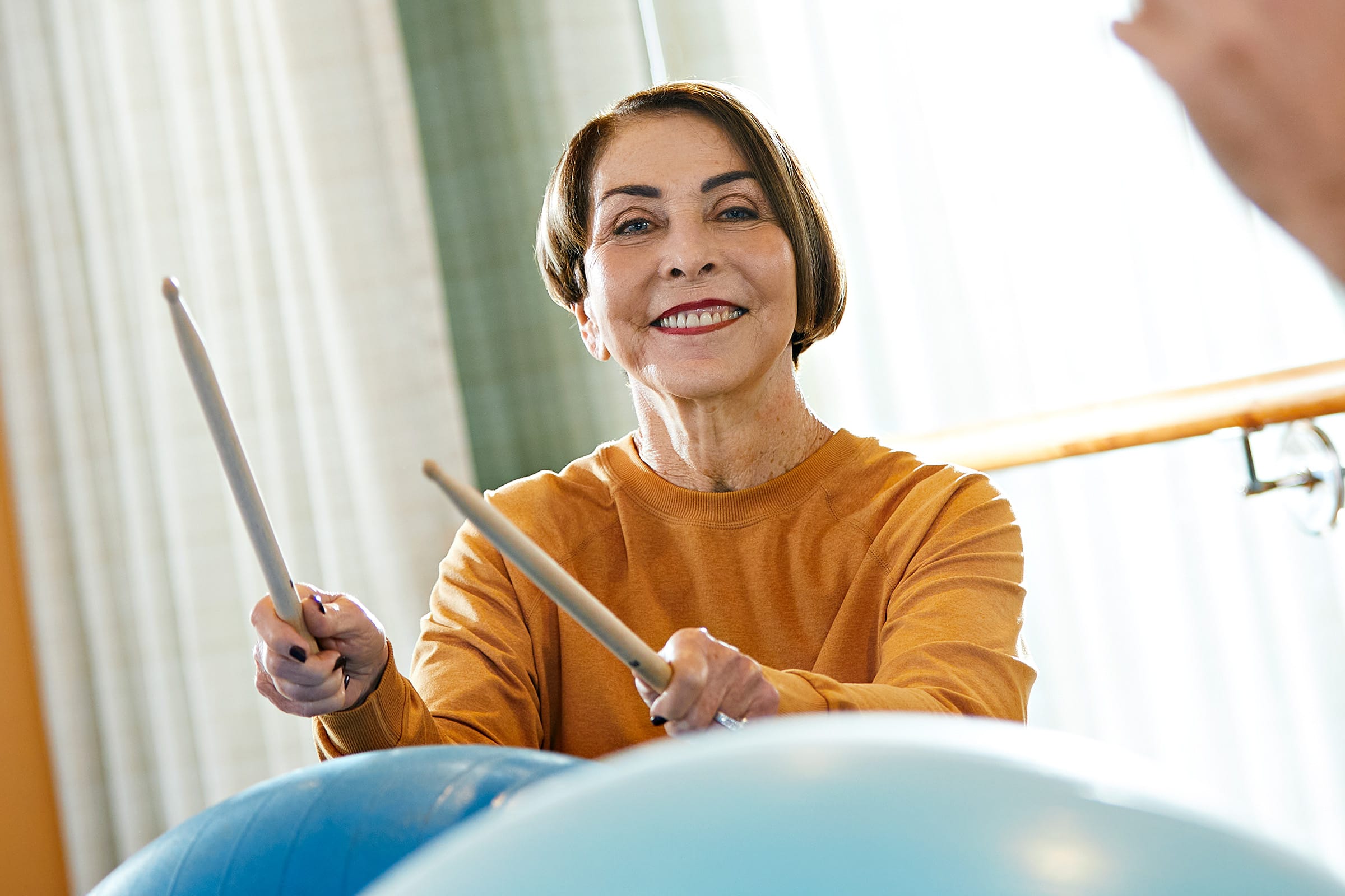 An older woman smiles while holding drumsticks and playing on large exercise balls in a bright room, suggesting a fun fitness or music activity.