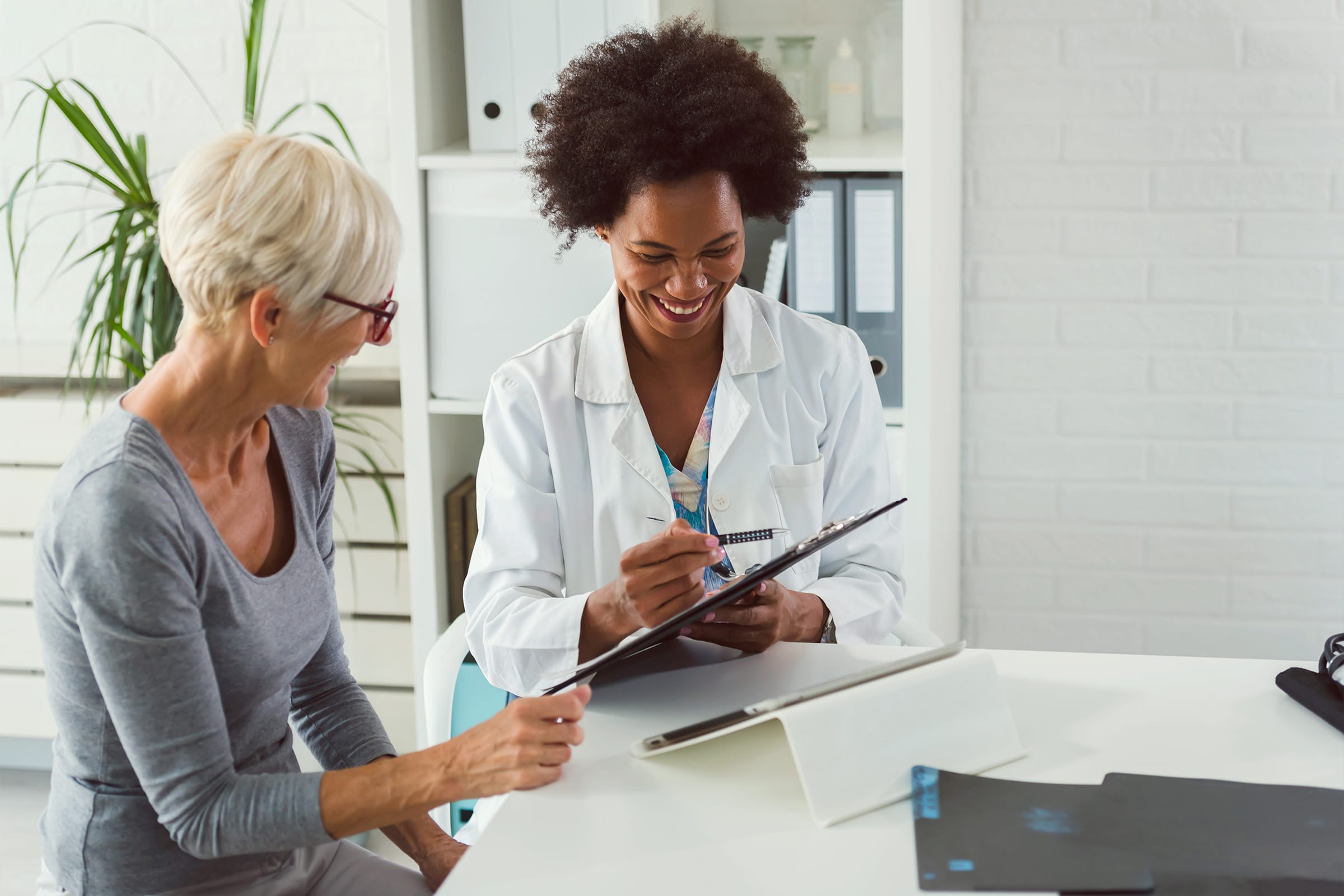 A doctor and an older woman sit at a desk, smiling and discussing paperwork. The doctor is wearing a white coat and holding a clipboard, while the woman is pointing at a document. Office shelves and a plant are visible in the background.