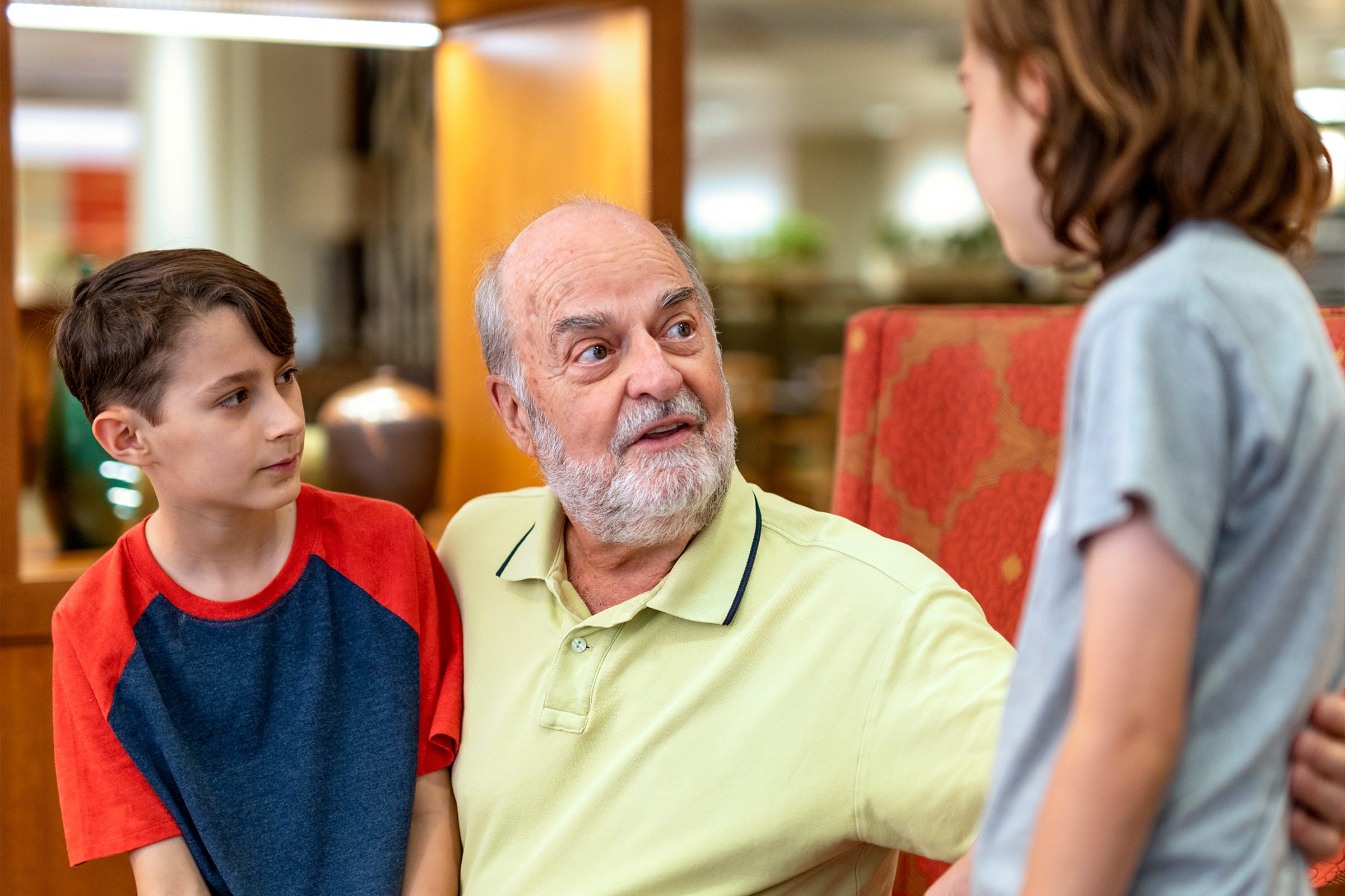 _photo-secure-future An older man with a white beard sits in a chair, talking to two children. One child sits beside him, while the other stands in front. They appear to be having an engaged, friendly conversation indoors.