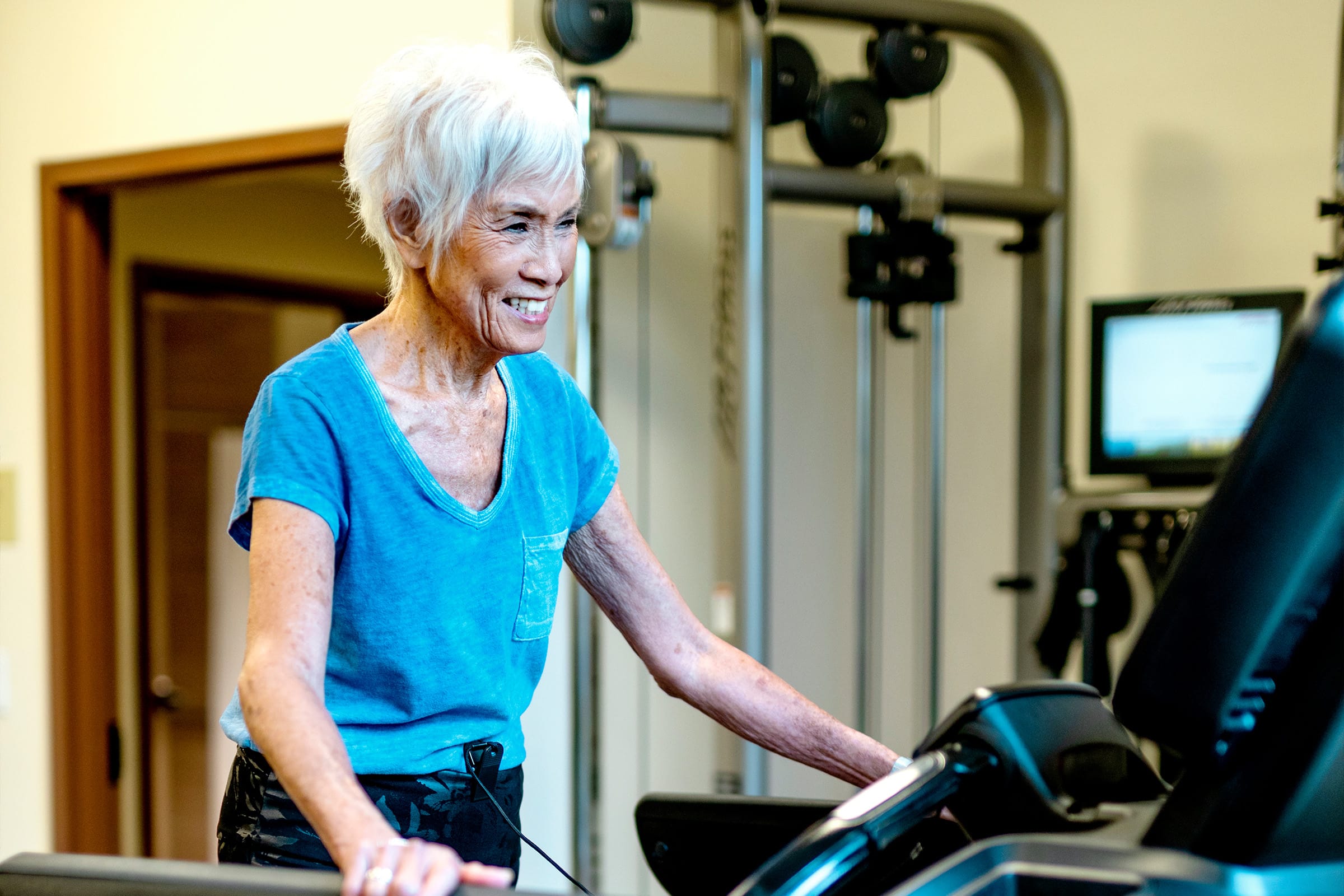 An older woman with short white hair and a blue T-shirt smiles while walking on a treadmill in a gym, with exercise equipment visible in the background.