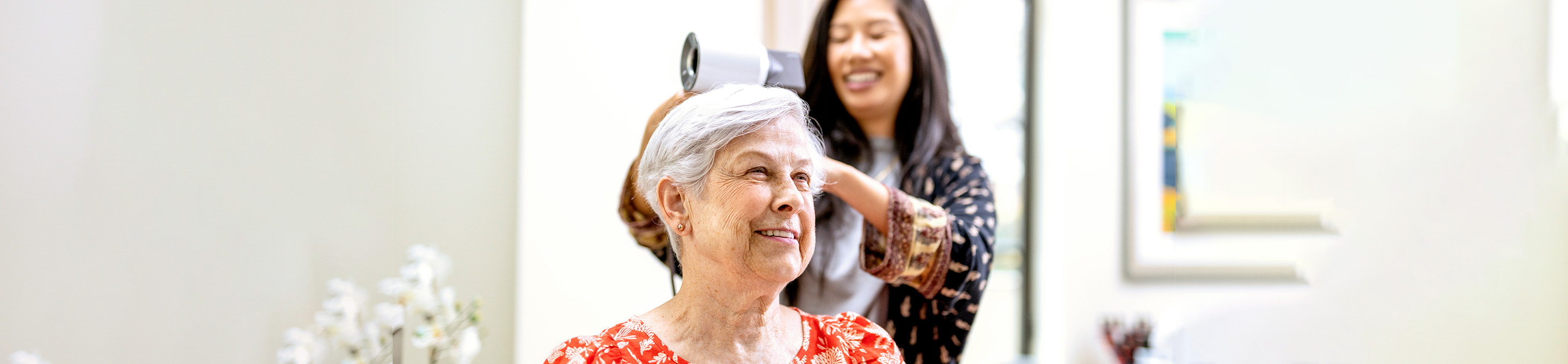 An older woman with white hair and a floral top smiles as a younger woman blow-dries her hair in a bright, modern salon.