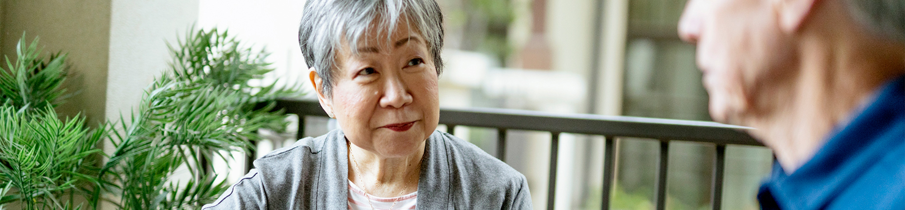 An older woman with short gray hair and a light gray sweater sits and listens attentively to another person, who is slightly out of focus, in an outdoor setting with greenery and a railing in the background.