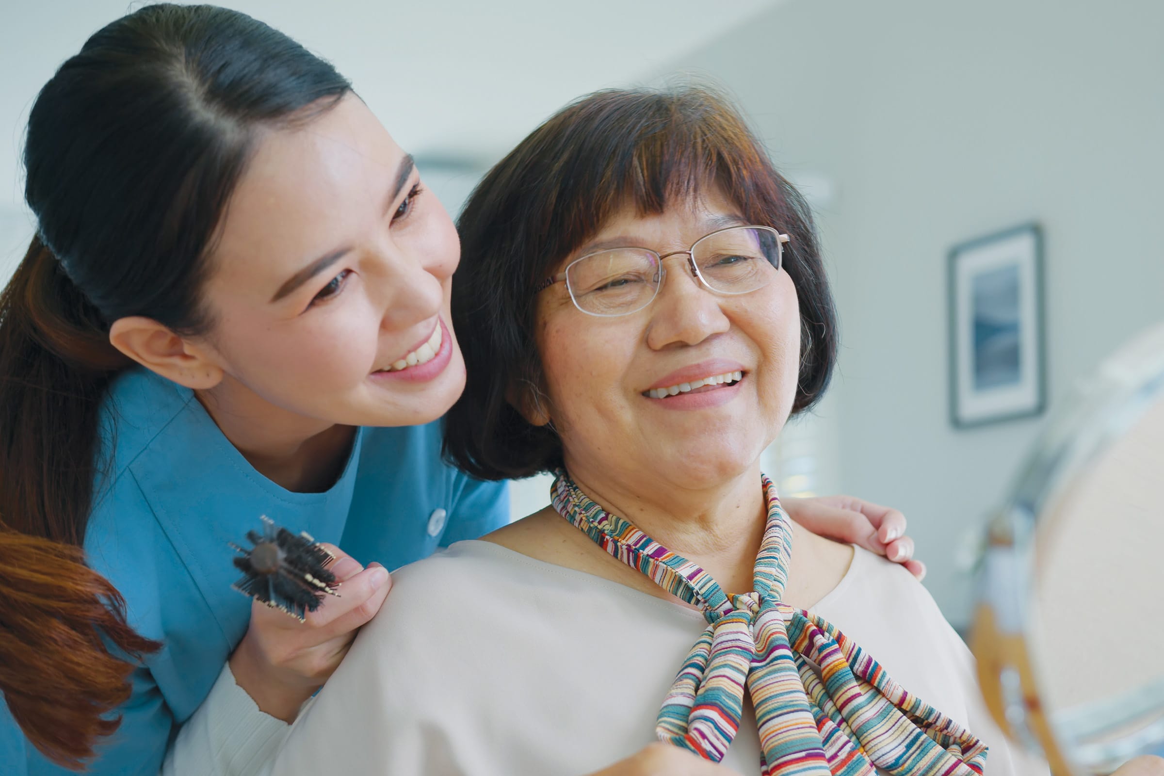 A young caregiver in a blue uniform smiles as she brushes the hair of an older woman with glasses and a colorful scarf, both appearing happy in a bright indoor setting.